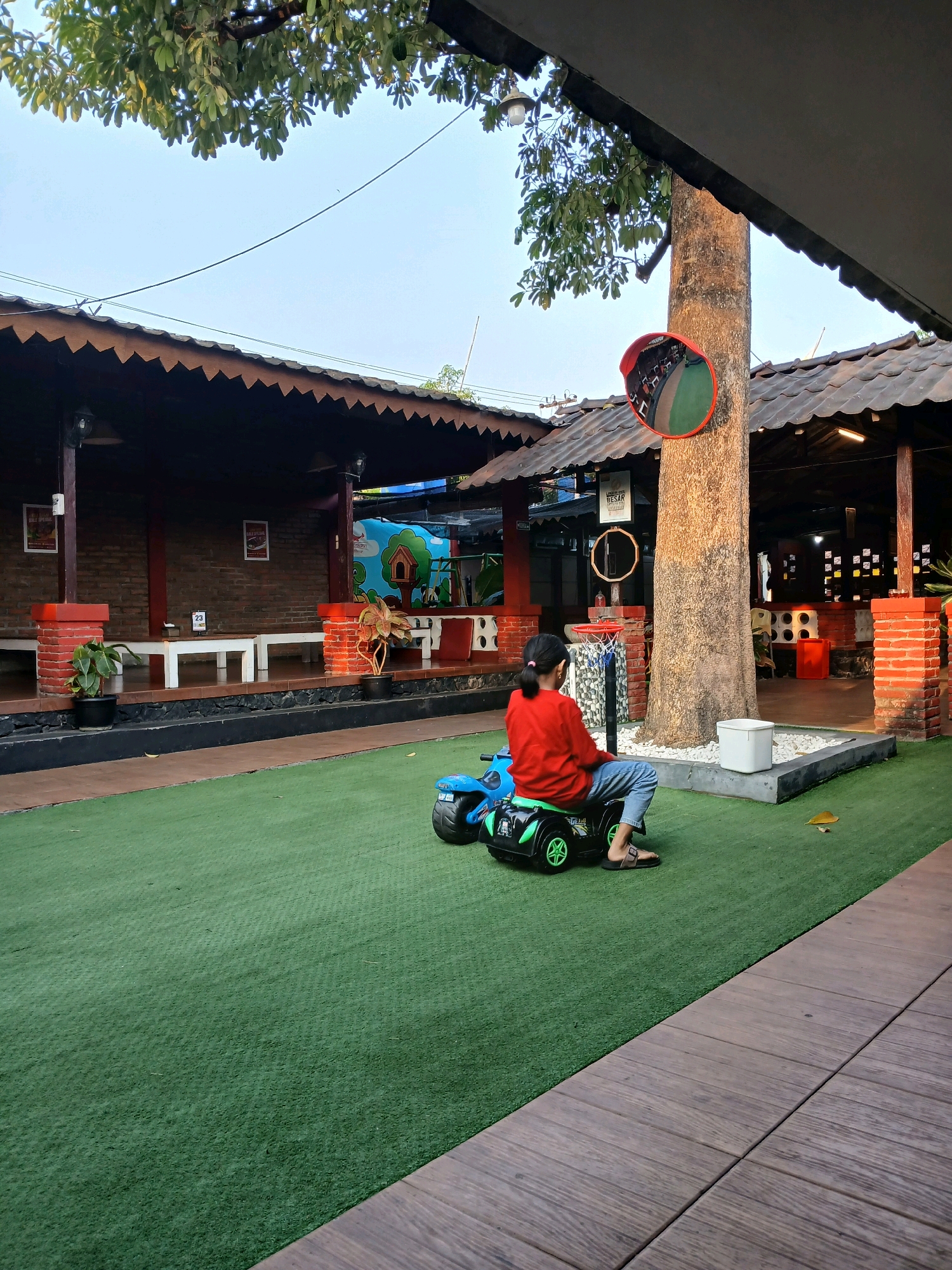 The image depicts an outdoor area with artificial turf and a wooden walkway. In the center, a young person wearing a red long-sleeved shirt and blue jeans sits on a small, black and green toy car. The person is facing away from the camera, looking towards a large tree trunk. The tree has a convex mirror attached to it. Behind the tree, a roofed structure with wooden beams and tiling is visible. Further back, there are brick walls, white benches, and potted plants. A basketball hoop and backboard are positioned near the tree. The overall scene suggests a recreational or dining area, possibly a cafe or a park in Yogyakarta, Indonesia, given the architectural style and vegetation. The lighting indicates it is either early morning or late afternoon.