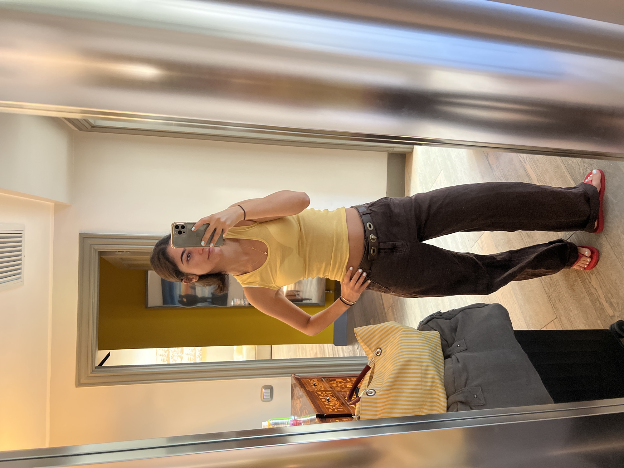 A young woman with dark brown hair stands in front of a mirror, taking a selfie with her phone. She is wearing a yellow tank top, dark brown wide-leg pants, a black belt, and red flip-flops. She has her right hand on her hip and her left hand holding her phone. In the background, a doorway leads to another room with yellow walls. There is a dresser with drawers to the left of the woman, and on top of it is a yellow and white striped tote bag. In front of the tote bag, there is a gray duffel bag, and below that, a black rolling suitcase. The floor is tiled with a light wood pattern. The scene appears to be inside a hotel room, possibly in Naples, Italy, given the style of the dresser and the warm lighting.