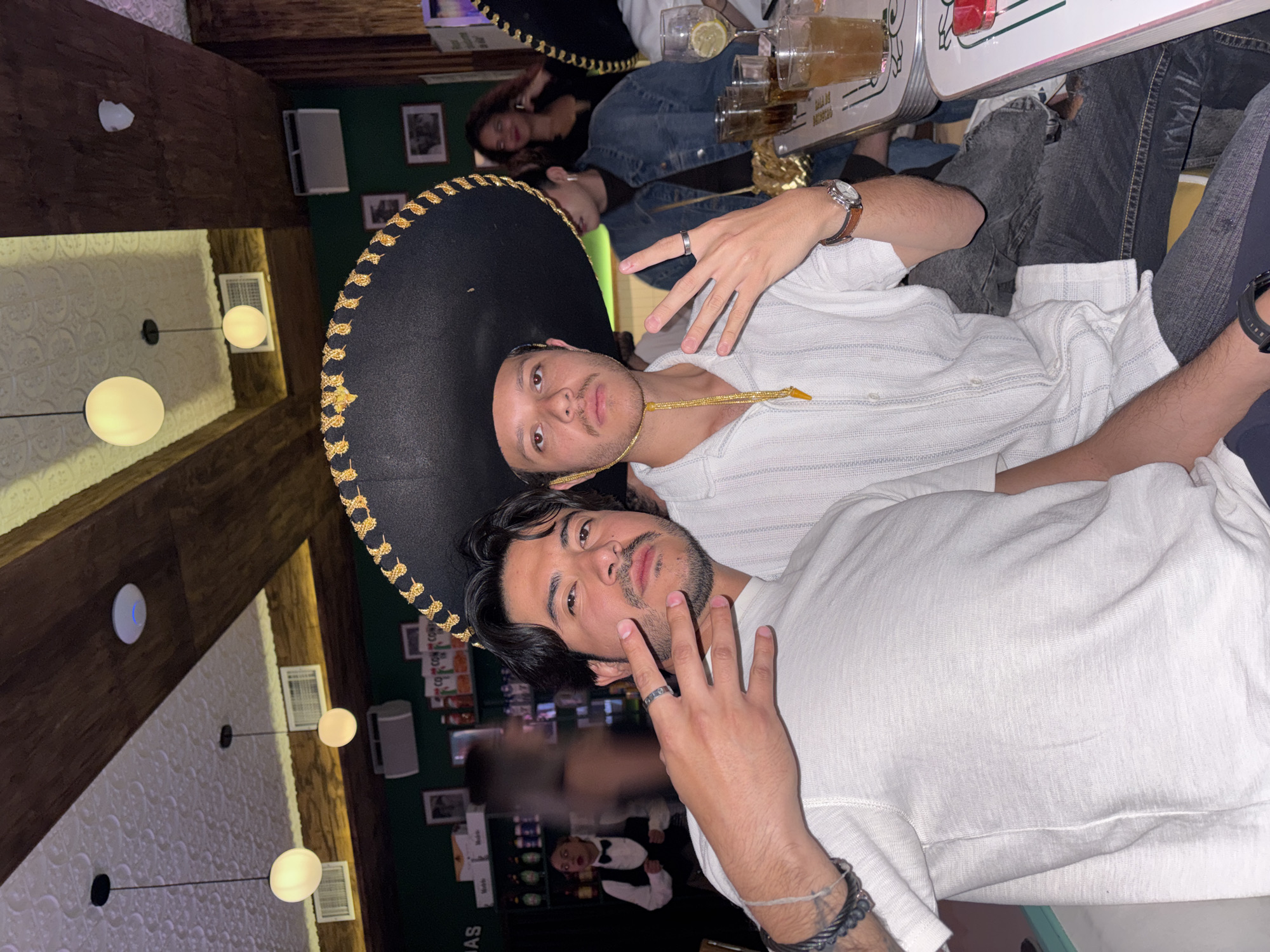 The image depicts two young men posing for a photo inside a bar or restaurant, possibly in Puebla, Mexico. The man on top is wearing a traditional black sombrero with gold trim and a white button-down shirt, he is holding up two fingers. The other man is wearing a white t-shirt and has a bracelet on his left wrist. There are drinks on the table. In the background, other patrons are visible, and the interior decor includes a green wall with framed pictures and a patterned ceiling with globe light fixtures. A waiter is seen in the background.