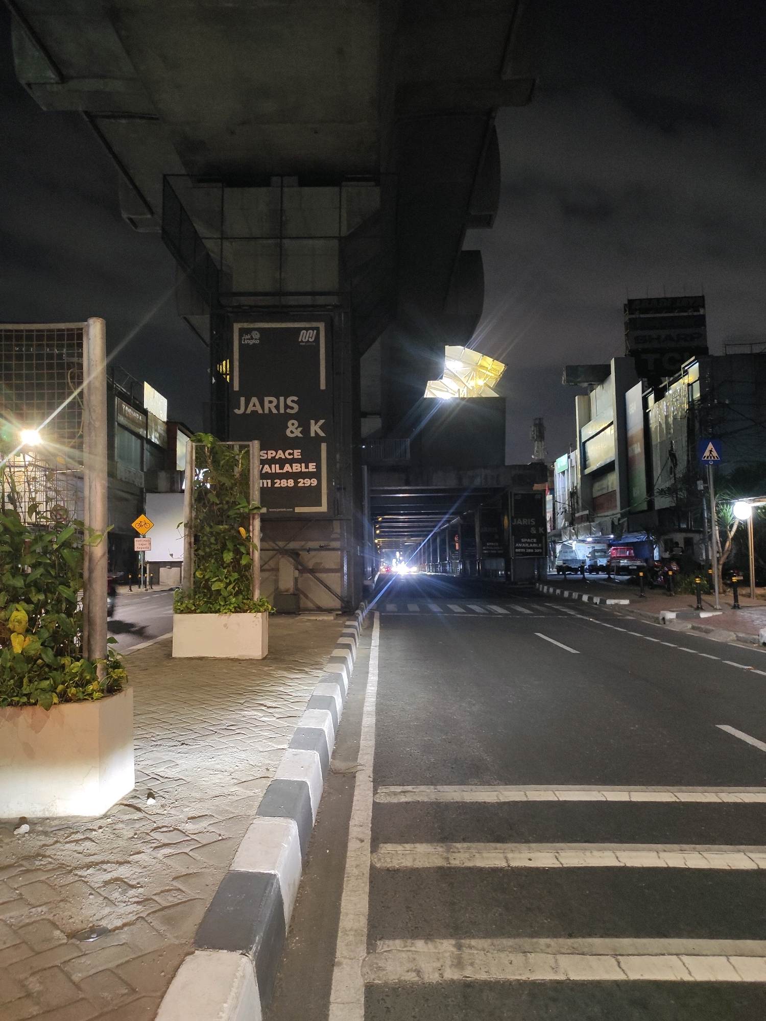 This image captures a nighttime street scene in South Jakarta, Indonesia, likely near an elevated infrastructure such as a highway or railway. The foreground features a pedestrian walkway with a checkered pattern of gray and white concrete curbs bordering a paved surface. A large rectangular planter with lush green foliage and yellow flowers sits on the left side of the walkway, illuminated by a bright lamp post to its left. To the right of the walkway is a dark asphalt road with clear white pedestrian crossing stripes.
In the mid-ground, beneath the concrete structure, a large black billboard displays "JARIS & K" in white lettering, with "SPACE AVAILABLE" and a phone number below it. Further down the road, vehicles are visible, their headlights creating streaks of light in the darkness. The road curves to the right and disappears into the distance.
The background is dominated by the imposing concrete support structures of the elevated infrastructure. Beyond these structures, several buildings are visible, some with illuminated windows and signage, including "SHARP" and "TOSHIBA". The sky is dark and cloudy, typical of nighttime. The overall atmosphere is one of an urban environment at night, with artificial lighting creating distinct contrasts.