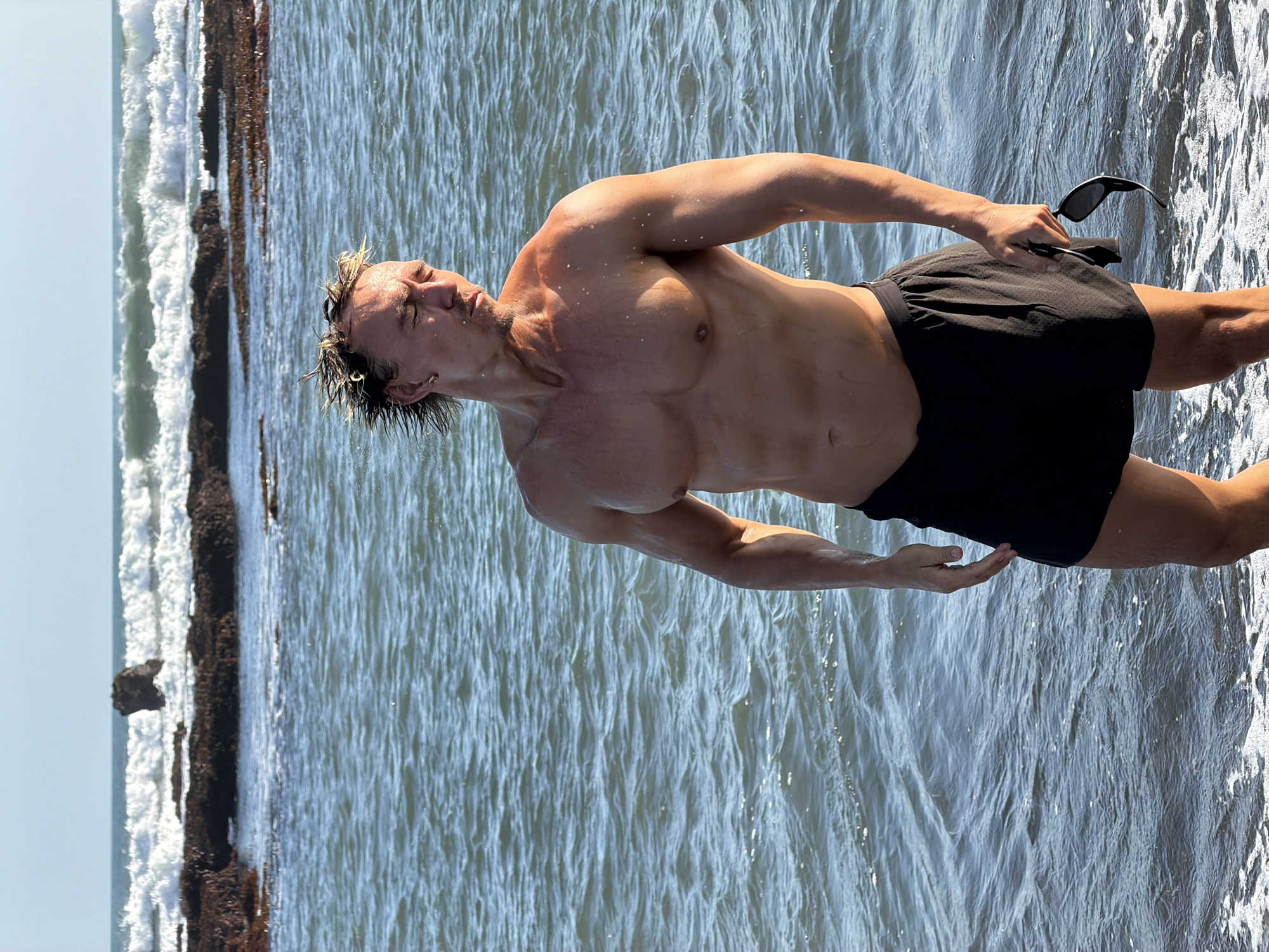 An athletic man stands in the shallow water at Batu Bolong Beach in Mengwi, Indonesia. He is shirtless and wearing black swim trunks, holding a pair of sunglasses in his right hand. Water droplets are visible on his skin and hair, suggesting he has recently been in the ocean. The ocean is calm with small waves breaking in the background, and rocky outcrops are visible in the distance. The sky is clear and blue.