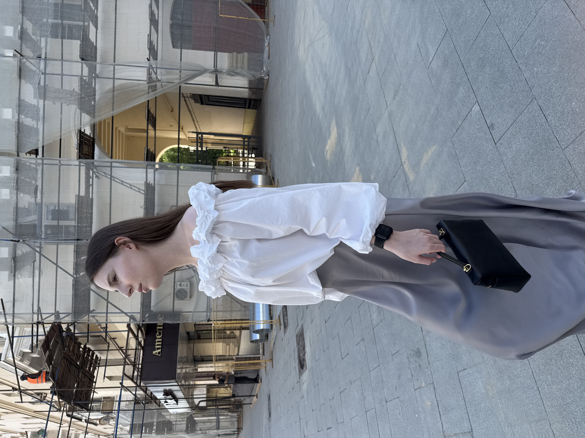 A young woman with long brown hair is captured in a profile view, walking along a city sidewalk. She is wearing an off-the-shoulder white blouse with ruffled trim and voluminous sleeves, paired with a long, flowing grey skirt. She carries a small black rectangular handbag with a top handle and gold hardware. A black smartwatch is visible on her left wrist. The background features a building undergoing renovation, with extensive scaffolding and protective sheeting covering its facade. A construction worker in an orange vest can be seen on a platform high up on the scaffolding. Yellow construction barriers are also present. The paving stones of the sidewalk are clearly visible. The scene appears to be set in an urban environment, likely Moscow, Russia, given the architectural style and the presence of construction typical of city development.