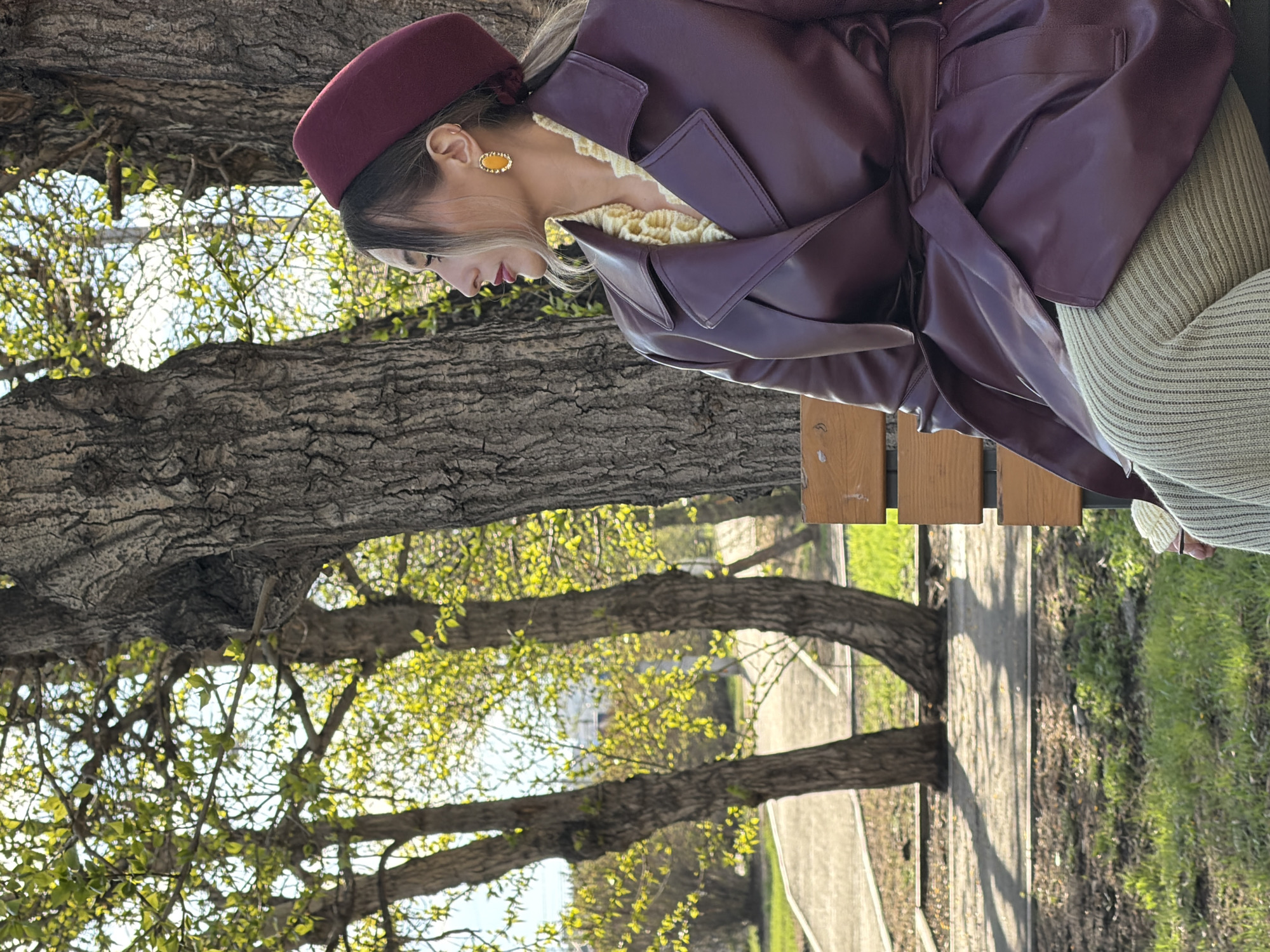 A young woman wearing a burgundy hat, a light yellow crocheted collar, and a purple leather-like coat is sitting on a wooden bench in a park-like setting. The park features mature trees with budding green leaves. A paved pathway is visible in the background. The photo appears to be taken outdoors in daylight. The location is Krasnoyarsk, Russia.
