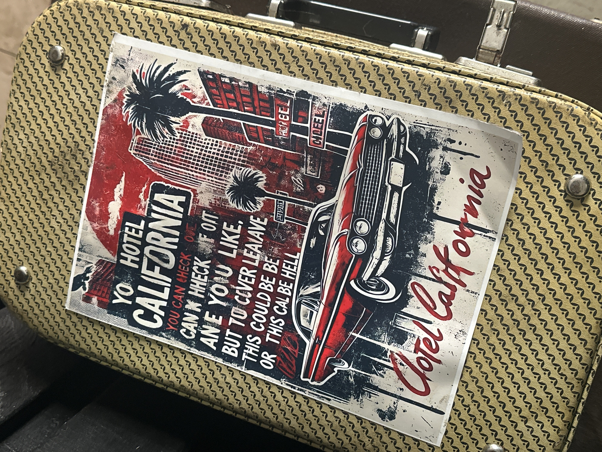 The image shows a close-up of a vintage suitcase covered with a sticker. The sticker is a design referencing the song "Hotel California." It features stylized palm trees, buildings, and a classic car, rendered in red, white, and black. The text on the sticker includes the phrase "Hotel California" and lyrics from the song. The suitcase itself has a textured, patterned surface. There is no indication that the photo was taken in Andreyevka, Russia, or Russia itself.