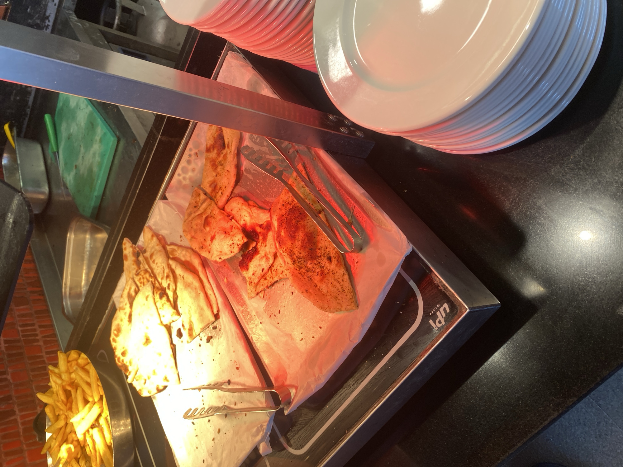 The image shows a close-up view of a food buffet in Aksu, Turkey, with a focus on a tray of flatbreads and a metal serving utensil. The flatbreads appear to be seasoned with spices and are arranged in a stack on a white paper liner atop a heated serving tray. To the left of the flatbreads, a bowl of french fries is visible. In the foreground, a stack of white plates is present. The overall lighting is warm, with a red hue illuminating the food. The background shows a brick wall and kitchen equipment.