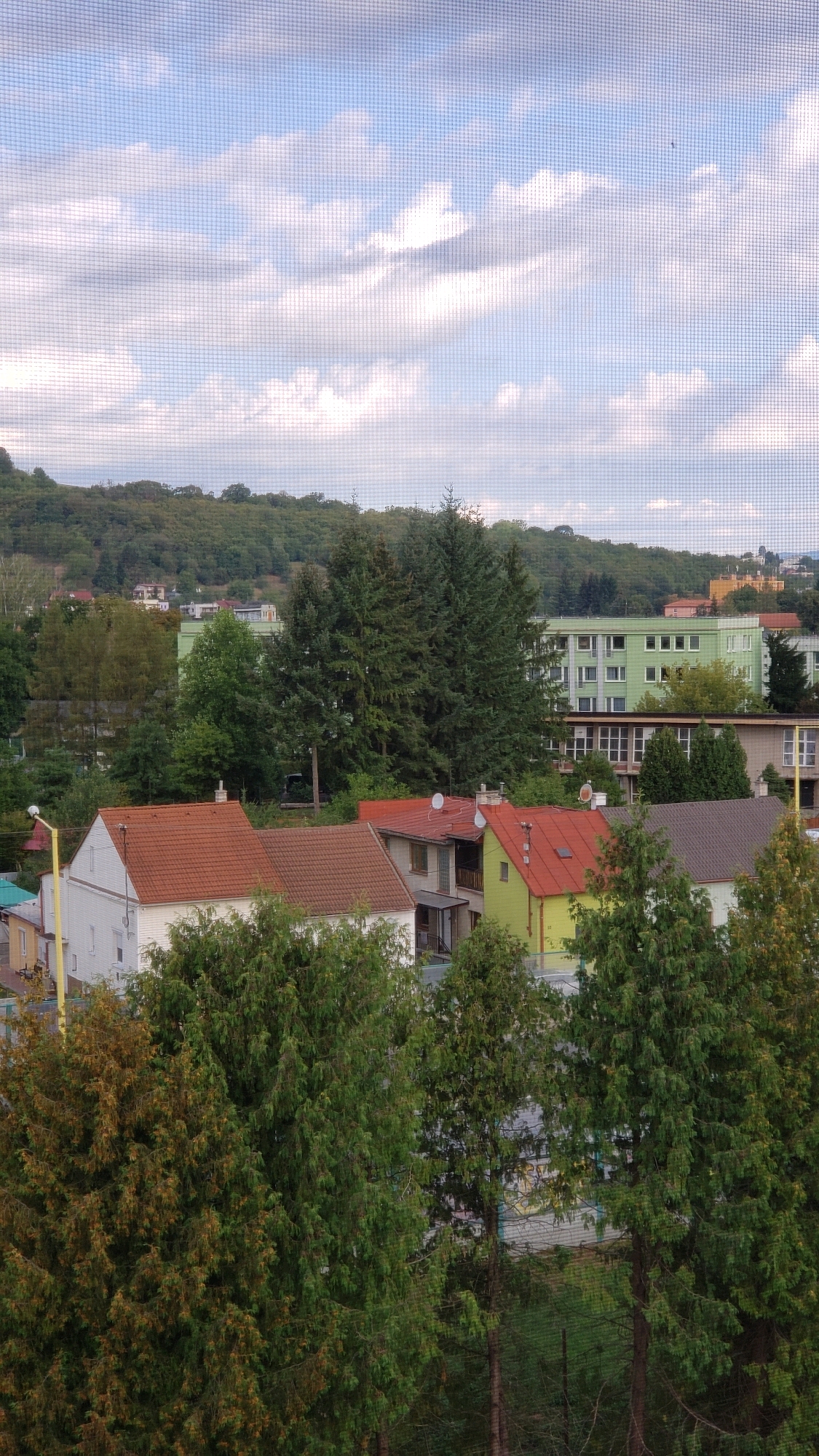 The image depicts a view of residential buildings and trees in the city of Prešov, Slovakia, seen through a window screen. The foreground is dominated by lush green trees, with one tree in the lower left having a slightly autumnal hue. Behind the trees, several houses with red tiled roofs are visible, along with buildings of varying architectural styles, including a prominent light green apartment building. In the background, a tree-covered hill rises, with more buildings scattered on its slopes. The sky is partly cloudy with a soft blue hue. The overall impression is of a suburban or semi-urban residential area.