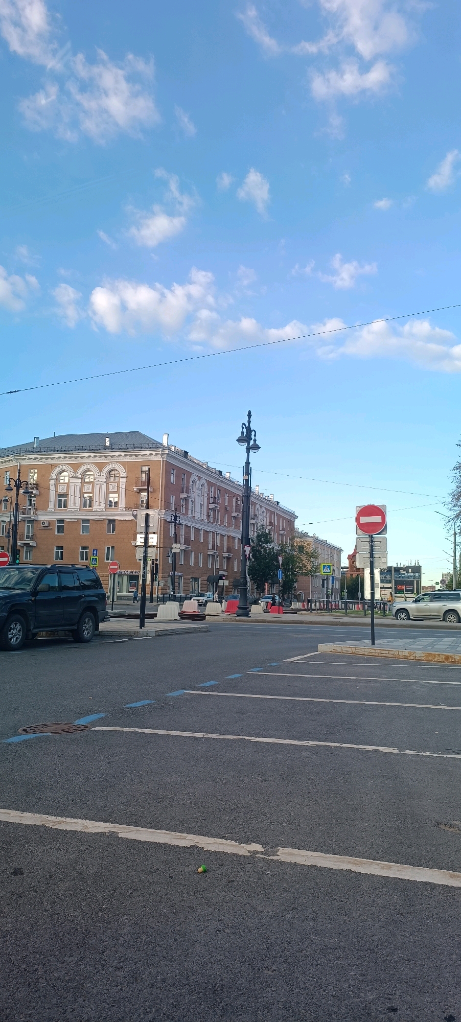 The image captures a view of a city street in Perm, Russia, with a large, multi-story building dominating the background. The building, appearing to be made of brick and adorned with architectural details, features numerous windows and a mansard roof. In the foreground, a paved road with lane markings is visible. A dark-colored SUV is parked on the left side of the street. Traffic signs, including a "No Entry" sign, are present along the road. Several streetlights with ornate designs are also seen. The sky is blue with scattered white clouds. The overall scene suggests a daytime urban setting with a mix of residential and commercial elements.