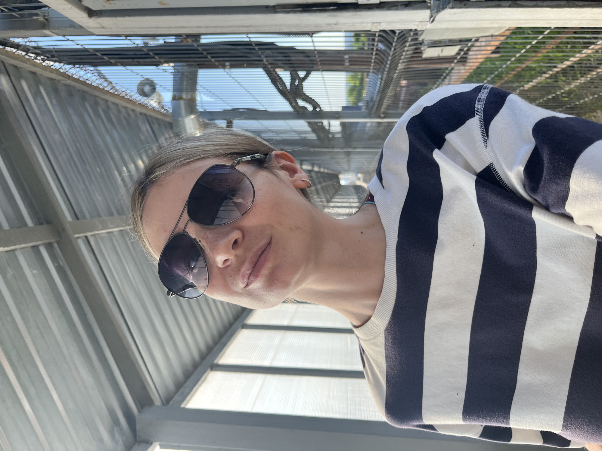 A woman wearing sunglasses and a black and white striped shirt is looking down at the camera. She is outdoors, possibly under a metal structure with a grated roof. The location is in the city of Chaikovsky, Russia.