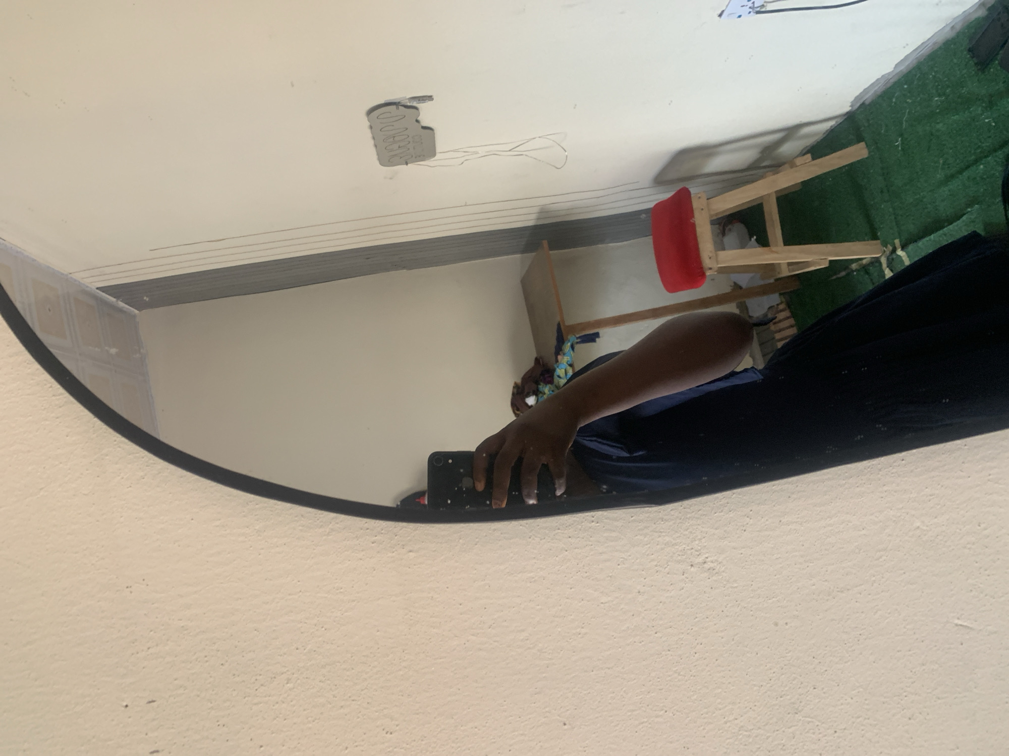 In Jauro Yinu, Nigeria, an image captures a dimly lit interior space with an oversized, oval mirror dominating the left side. A person, partially obscured by the mirror's reflection, stands holding a black smartphone, taking a selfie. They are wearing a dark blue garment. The reflected background reveals a textured, light-colored wall, a wooden stool with a red cushion, a small wooden table, and a white electrical outlet with wires. The floor is covered with a green carpet.