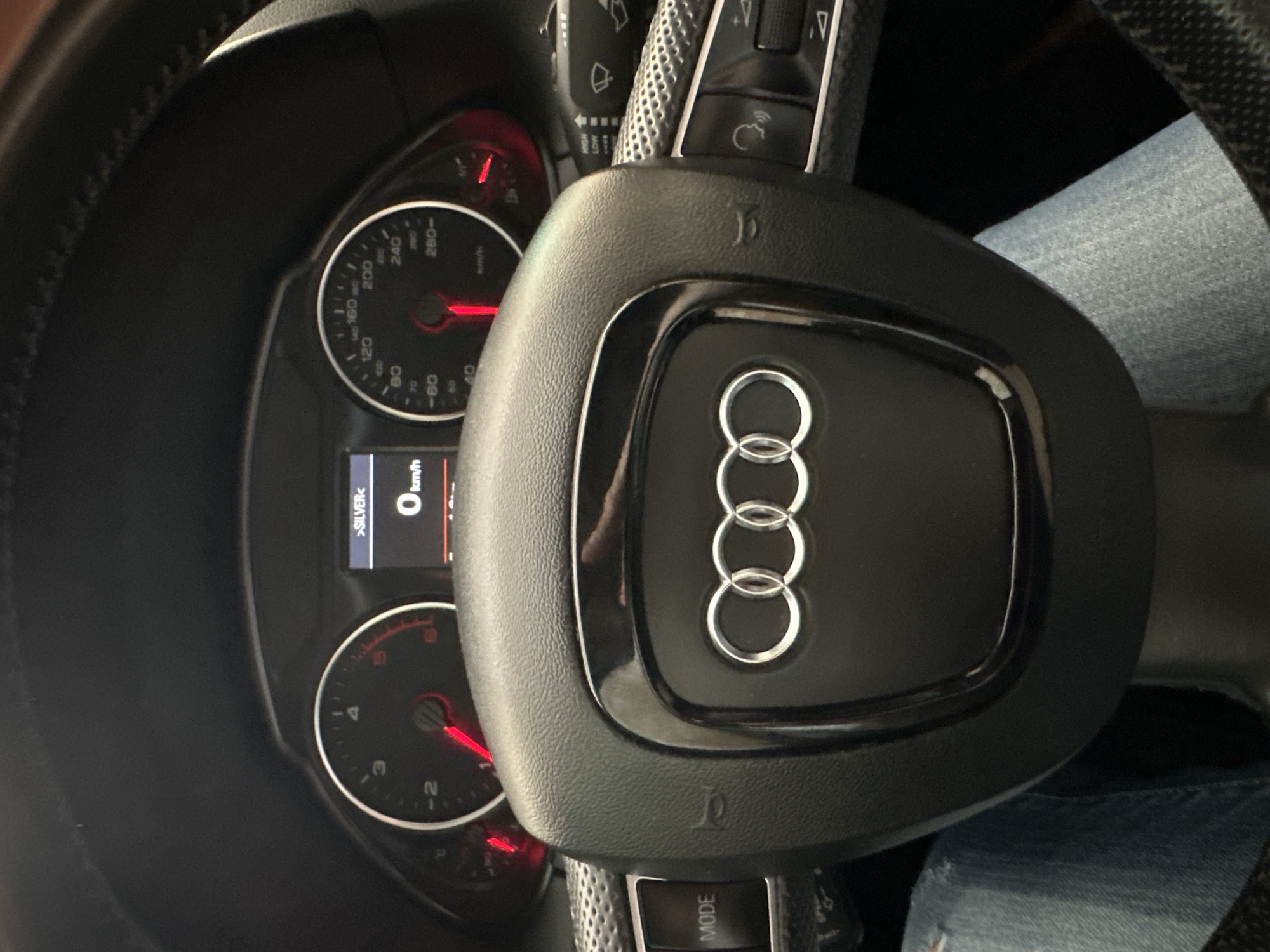 A close-up interior shot shows the dashboard and steering wheel of an Audi vehicle. The dashboard features circular gauges with red pointers, and a digital display reads "0 km/h" and ">SILVER<". The steering wheel is black with silver accents and has the Audi logo prominently displayed in the center. A small portion of a person's jeans-clad leg is visible to the right of the steering wheel. The image composition is focused on the vehicle's controls and instrumentation.