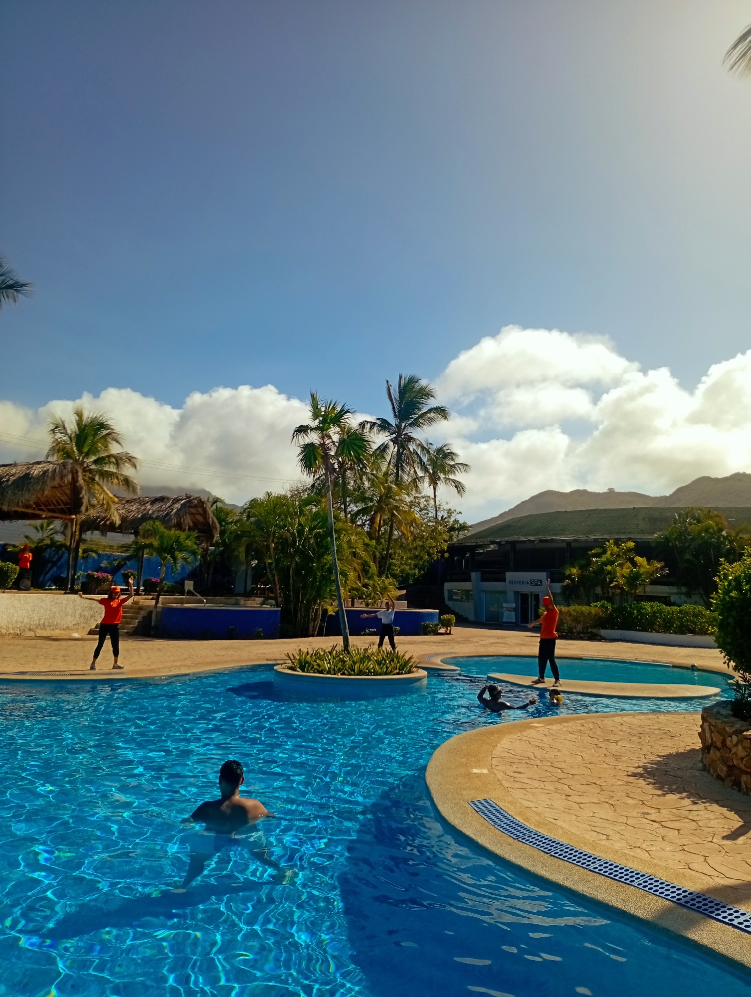 The image shows an outdoor swimming pool complex at a resort in Playa El Agua, Venezuela, under a partly cloudy sky.
The pool features an irregular shape with several connected smaller pools and islands of landscaping. The water is clear blue. A man is seen swimming in the pool, his back to the camera. Two people are visible in the adjacent pool, one appearing to be submerged with their head partially out of the water.
Around the pools, there are tan tiled walkways with blue drainage strips. Three people wearing orange shirts and dark pants stand near the pool's edge, two with arms raised. These appear to be instructors or staff leading an activity.
In the background, there are palm trees, thatched-roof structures, and buildings, possibly a resort or hotel complex. Hills are visible in the distance. The lighting suggests it is daytime with a bright sun.