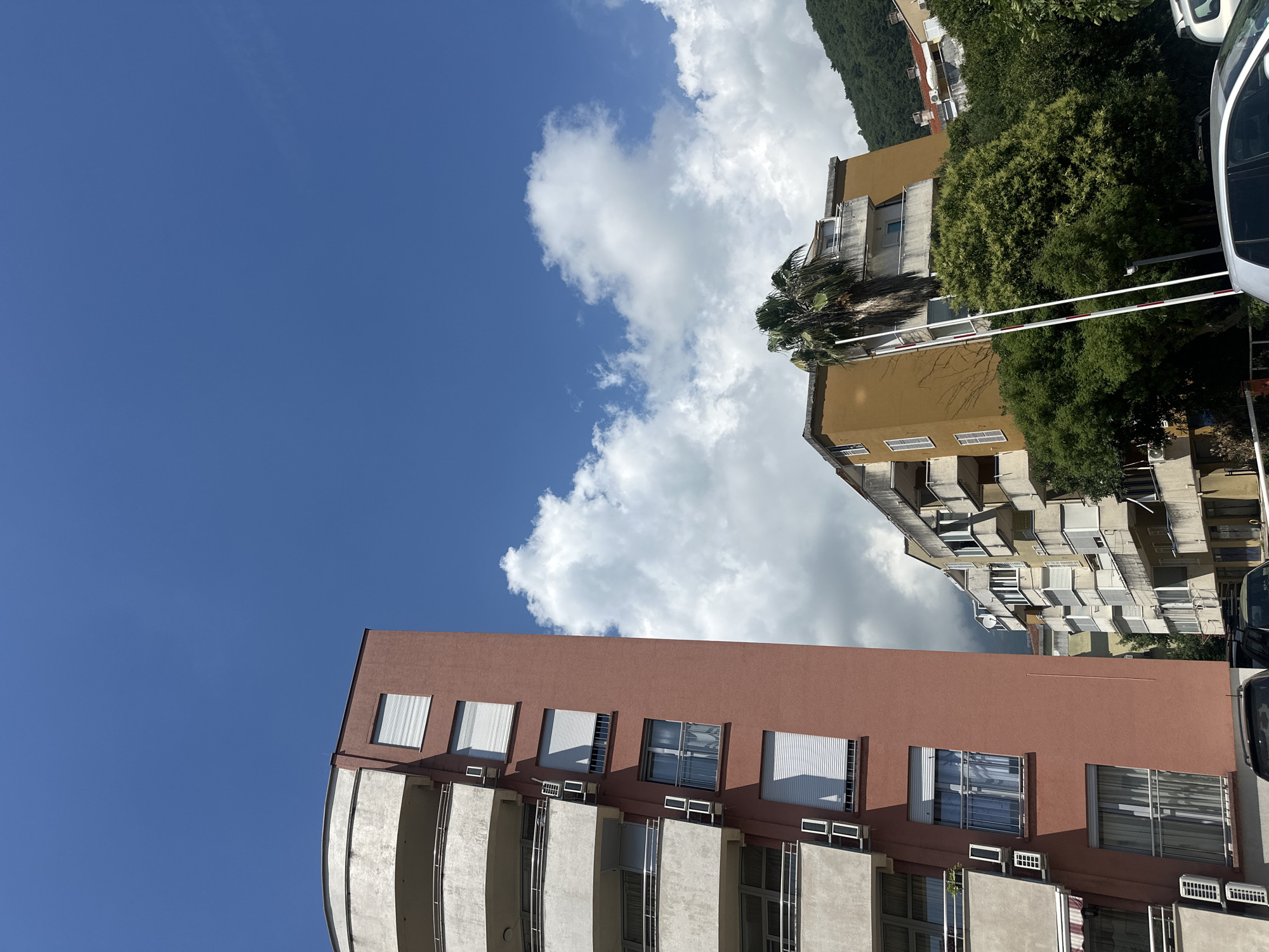 The image, captured during the day in Budva, Montenegro, displays a residential urban scene under a bright sky with scattered white clouds. A prominent multi-story apartment building occupies the left side of the foreground. This building has a reddish-brown upper section and lighter, possibly concrete, lower parts with a series of balconies. Its numerous windows feature white external blinds or shutters, and several small air conditioning units are mounted below them. A distinct curved architectural element is visible on the building's left side.
Further back and to the right, other multi-story apartment blocks with yellowish facades and numerous balconies are visible, interspersed with abundant green trees and vegetation, including a tall palm tree. In the distant background, densely forested green hills or mountains rise.
A white and red striped barrier arm is present in the mid-ground, suggesting an entrance or parking control. Fragments of vehicles, including a white car, are visible along the far right edge of the frame. The strong sunlight casts clear shadows, indicating a sunny daytime. No specific human activity or visible text is present, depicting a tranquil residential area typical of a Mediterranean coastal town.