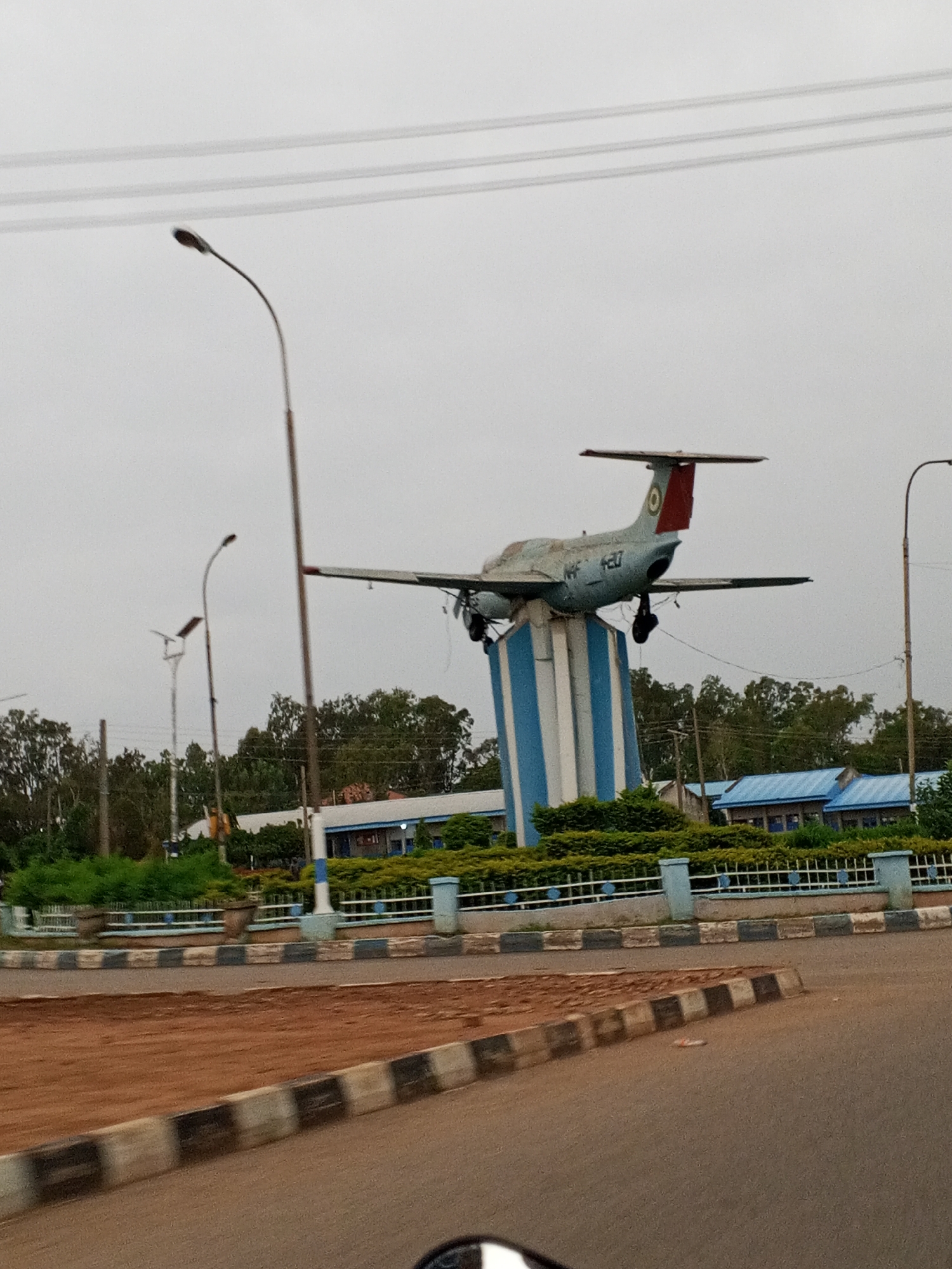 The image captures an outdoor scene in Barakin Lerawi, Nigeria, dominated by a monument featuring a vintage jet airplane displayed atop a cylindrical pedestal. The pedestal is painted with vertical blue and white stripes. The airplane itself is a light blue color, with red accents on its tail and some markings, including "420" and "NF" visible on the fuselage. The monument is situated in an urban or semi-urban environment, indicated by the presence of paved roads, streetlights, and buildings in the background. The buildings appear to be governmental or institutional, with blue roofs and light-colored walls. Lush green bushes and trees form a backdrop and surround the monument. The sky is overcast, suggesting a cloudy day. The foreground shows a section of a road and a sidewalk, with decorative curbing. The overall impression is one of a public display or landmark.