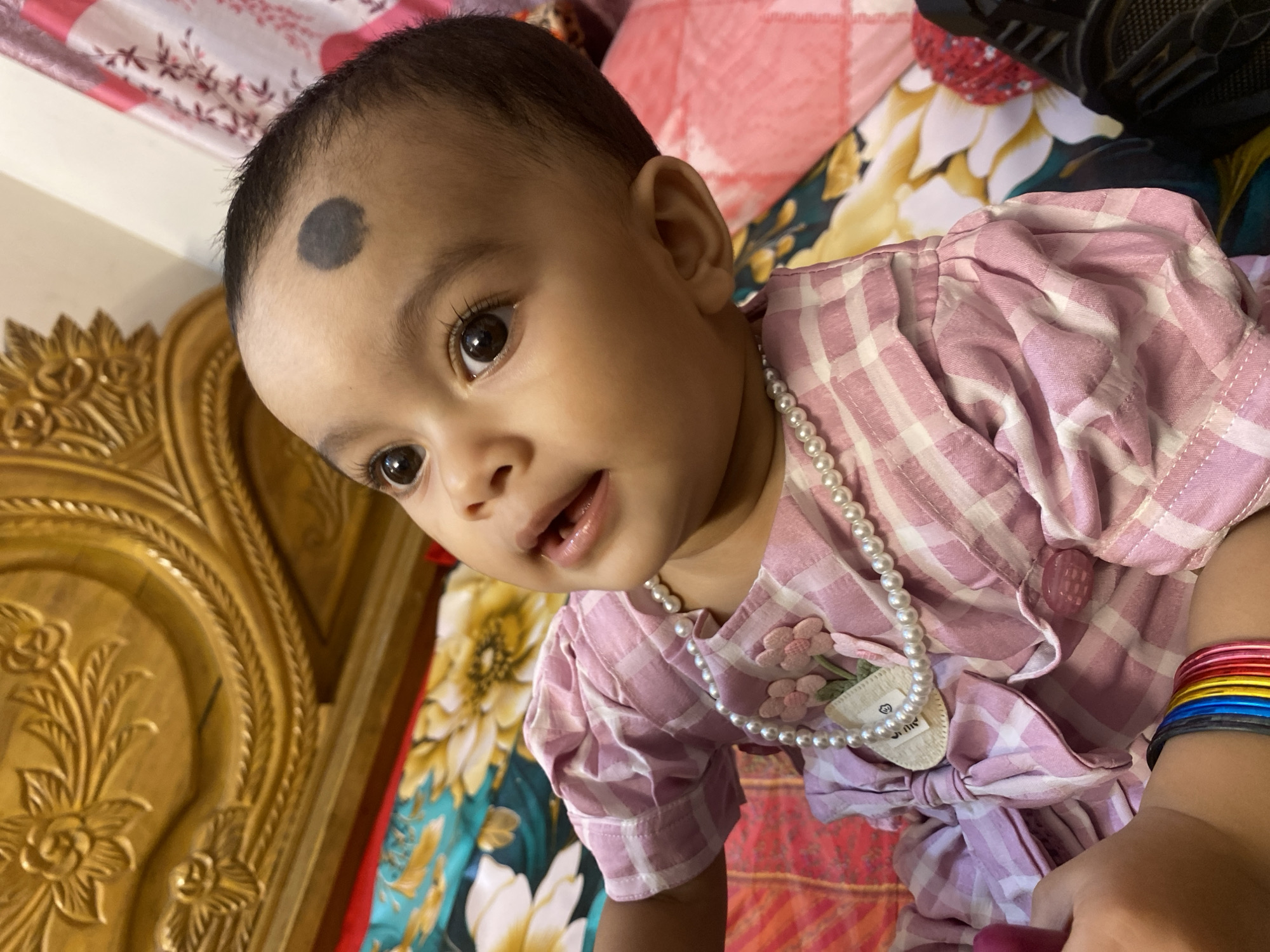 A close-up shot captures a South Asian baby girl wearing a pink and white checkered dress and a pearl necklace, sitting on a bed. The baby has a dark birthmark on her forehead. The background features a wooden headboard and patterned bedding, suggesting a domestic indoor setting. The image appears to be taken in Dhaka, Bangladesh, a city known for its vibrant culture and dense population.