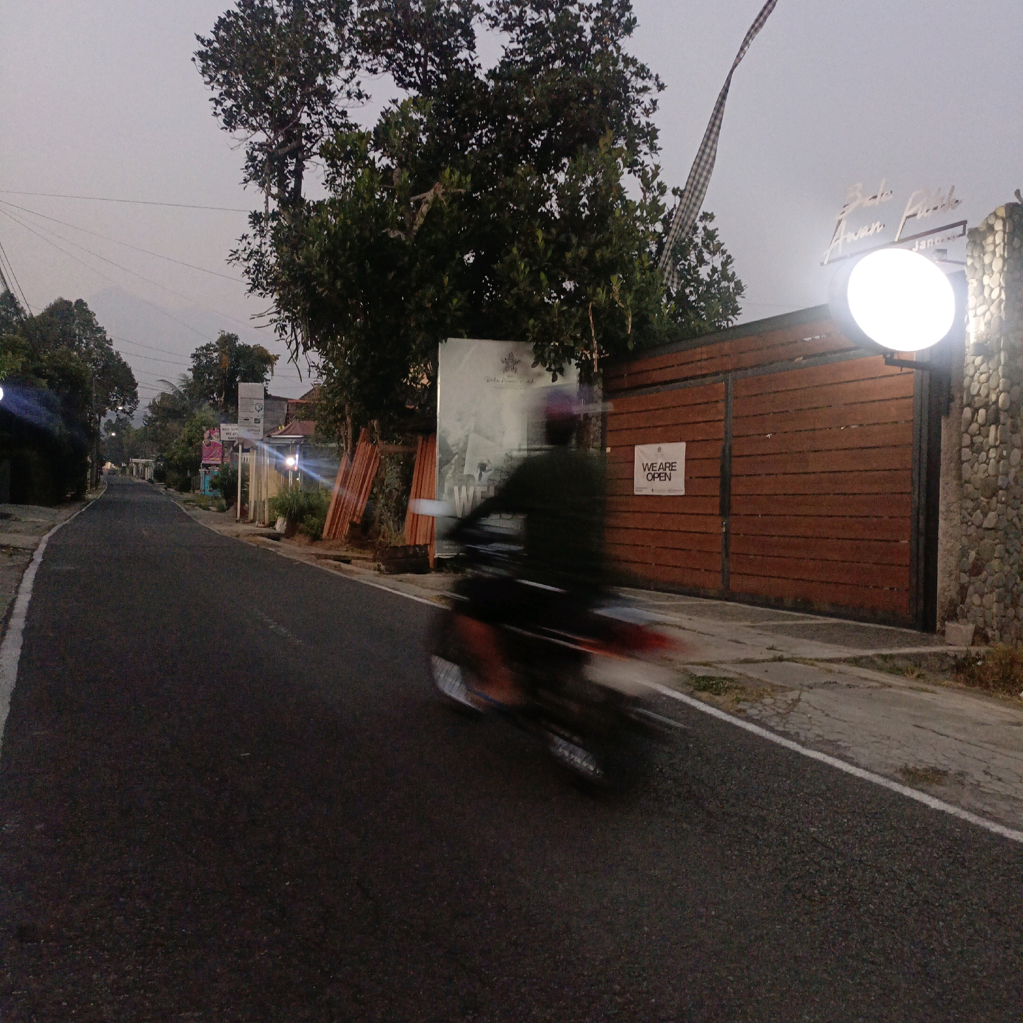 The image depicts a street scene in Salatiga, Indonesia, at dusk or early morning. A motorcycle is captured in motion, creating a motion blur effect, with its rider visible. The road is paved and has a white line marking its edge. On the right side of the road, there is a large wooden gate with a stone pillar. A circular light fixture illuminates the area in front of the gate, and a sign that reads "WE ARE OPEN" is affixed to the gate. Behind the gate, a large advertisement or poster is visible, partially obscured by trees and a banner flag. The sky is overcast, and trees line the street, with some buildings visible in the background. The overall impression is of a quiet street with an establishment that is open for business.