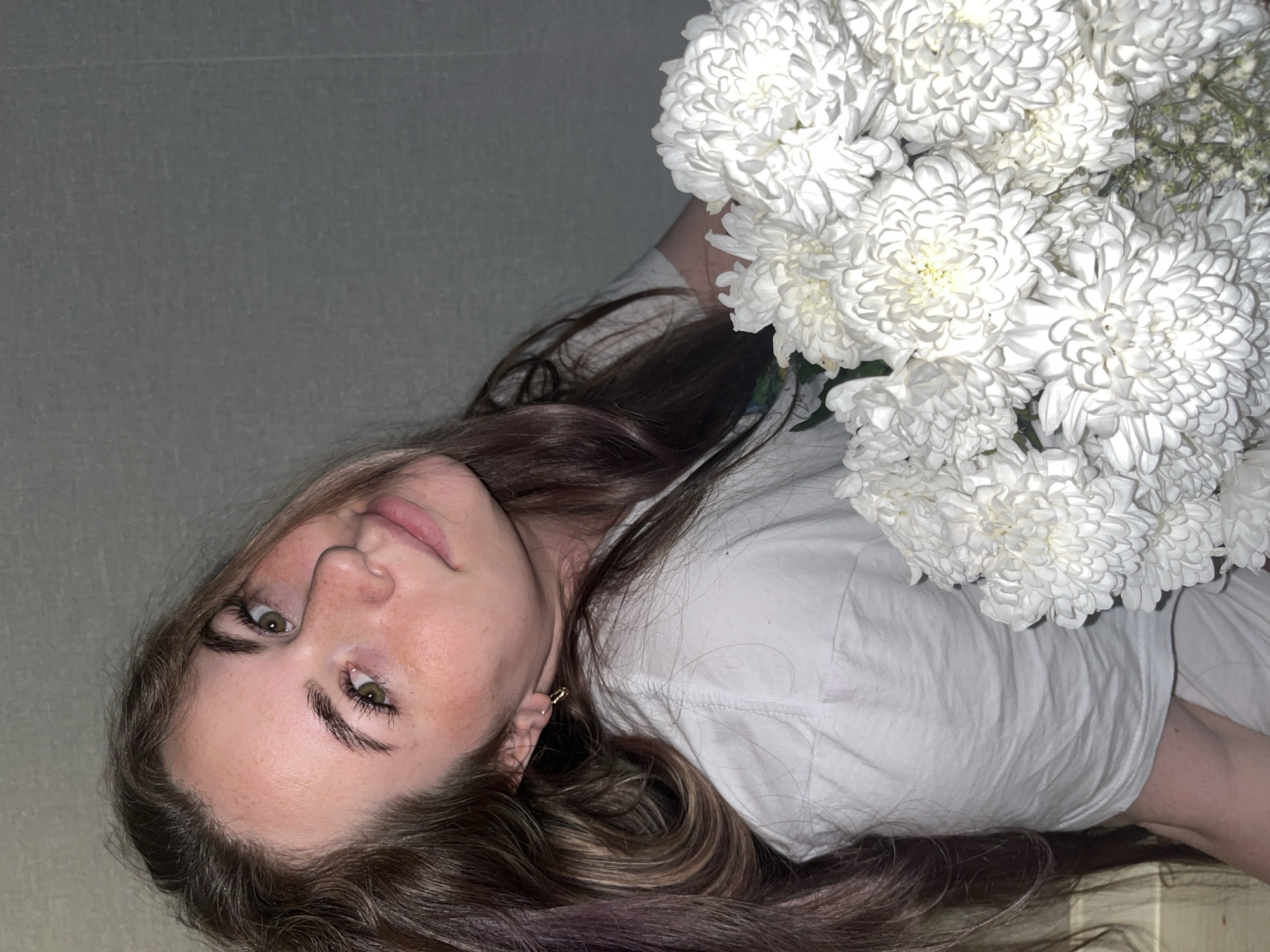 A young woman with long brown hair, fair skin, and freckles poses with a bouquet of white chrysanthemums. She wears a white shirt and gold hoop earrings. The background is a plain gray wall. There is no indication of this image being taken in Chaikovsky, Russia.