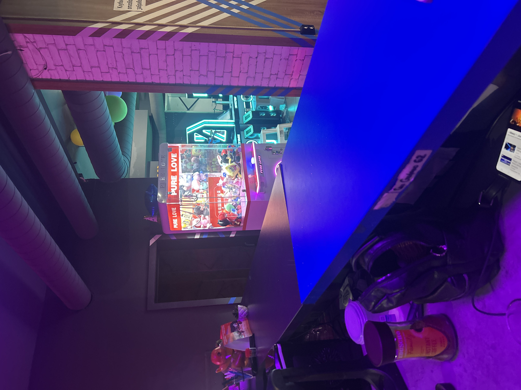 This indoor shot taken in Burgas, Bulgaria features an array of objects in a room illuminated with purple and blue lighting. In the left background are ventilation ducts with yellow and green balloons attached. Next to it is a claw machine filled with plush toys, labeled "PURE LOVE" in both English and Mandarin Chinese. The machine is illuminated with neon lights and is positioned near what appears to be a neon-lit arcade or recreational area.
In the foreground on the right, a desk or countertop is prominently featured, cluttered with items including an open black leather bag, a smartphone, a container with a purple lid, and a jar labeled "Conscito, INSTANT COFFEE." Various other items are situated on the lower left, including what appears to be a red figurine. The room has a playful, urban vibe, with the lighting adding to the modern and somewhat chaotic setting. There are no visible people in the shot.