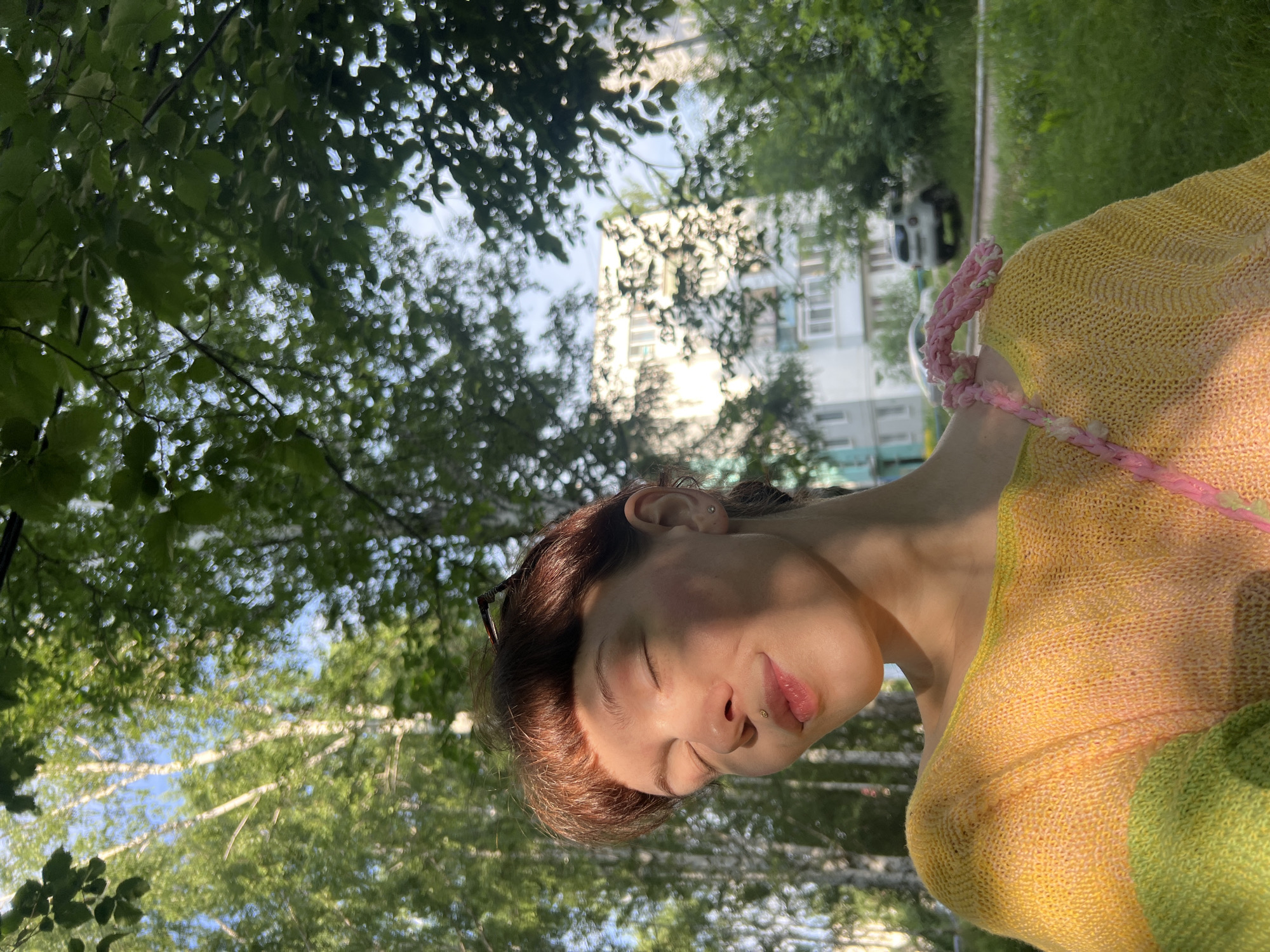 The image shows a young person with short reddish-brown hair and closed eyes, basking in sunlight. They are wearing a yellow knitted sweater with green accents and a pink braided strap. A subtle lip ring and a small stud earring are visible. The background features trees, a building, and a car, suggesting an outdoor urban setting. The dappled sunlight filtering through the leaves creates a serene atmosphere. The presence of birch trees in the background indicates a likely location in a temperate climate, and the architectural style of the building is consistent with Soviet-era housing found in many Russian cities, including Chaikovsky.