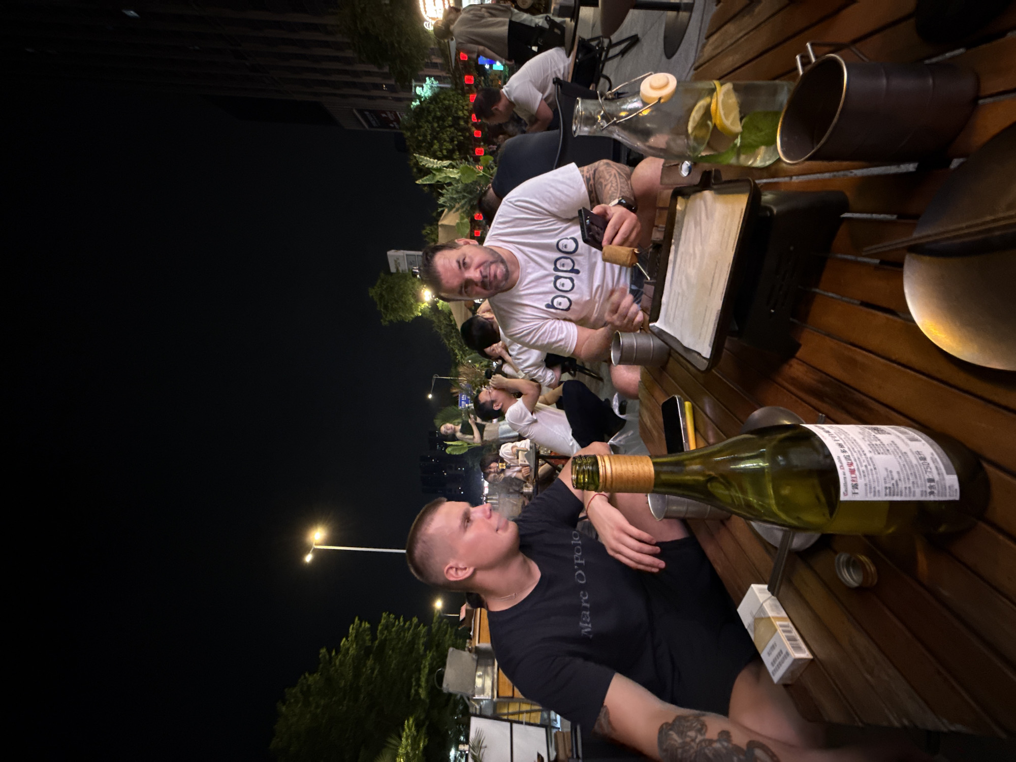 Two men are seated at a wooden table outdoors at night, in what appears to be a restaurant or bar area. The man on the left is wearing a black t-shirt with "Marc O'Polo" written on it and has short blond hair. He is looking to his right. The man on the right is wearing a white t-shirt with "bape" printed on it and has dark hair and a beard. He is holding a mobile phone in his left hand and a skewer with food in his right hand, looking towards the camera.
On the table in front of them are a green bottle of wine, a pack of cigarettes, a mobile phone, a glass bottle with a stopper filled with water and lemon slices and mint leaves, and a dark metal mug. A small grill with a sheet of white paper on top is also visible.
In the background, other people are seated at tables, and there are trees, streetlights, and buildings, suggesting an urban setting. The overall lighting indicates it is evening. The scene is consistent with an outdoor dining or social gathering in Haizhu District, China.