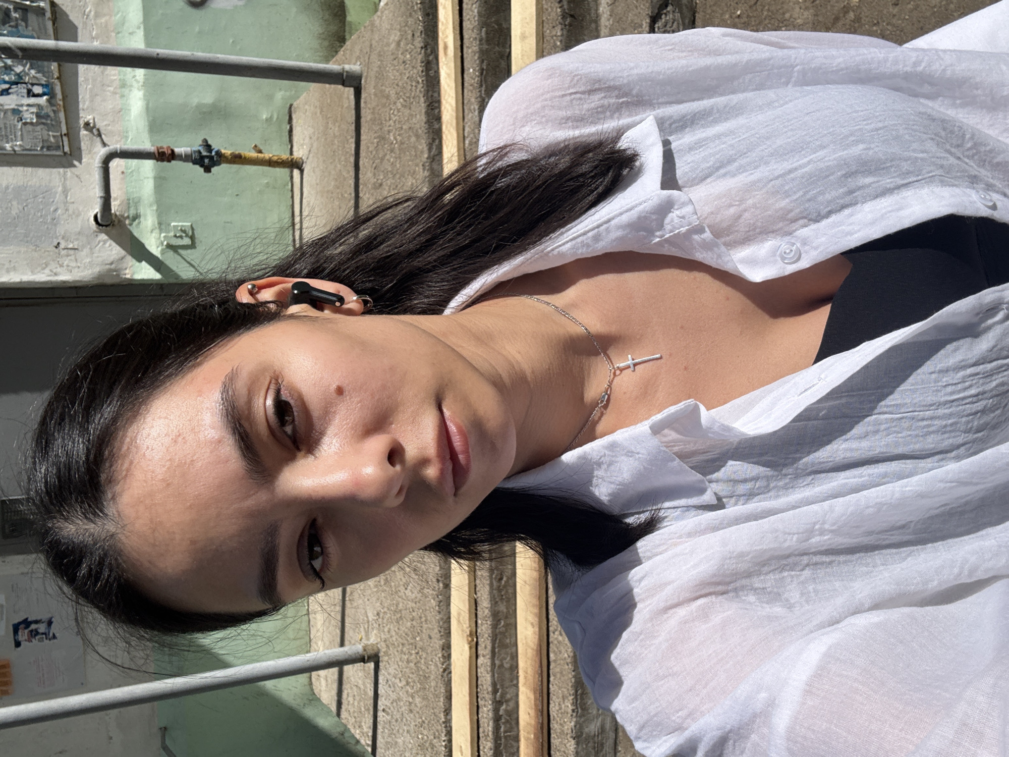 A woman is pictured outdoors, her face tilted towards the camera. She has dark hair and a silver necklace with a cross pendant. She wears a black top under a white, unbuttoned shirt. In her ear, there appears to be a dark-colored earbud or hearing aid. The background shows a concrete structure with a metal railing and pipes. The scene is situated in Chaikovsky, Russia.