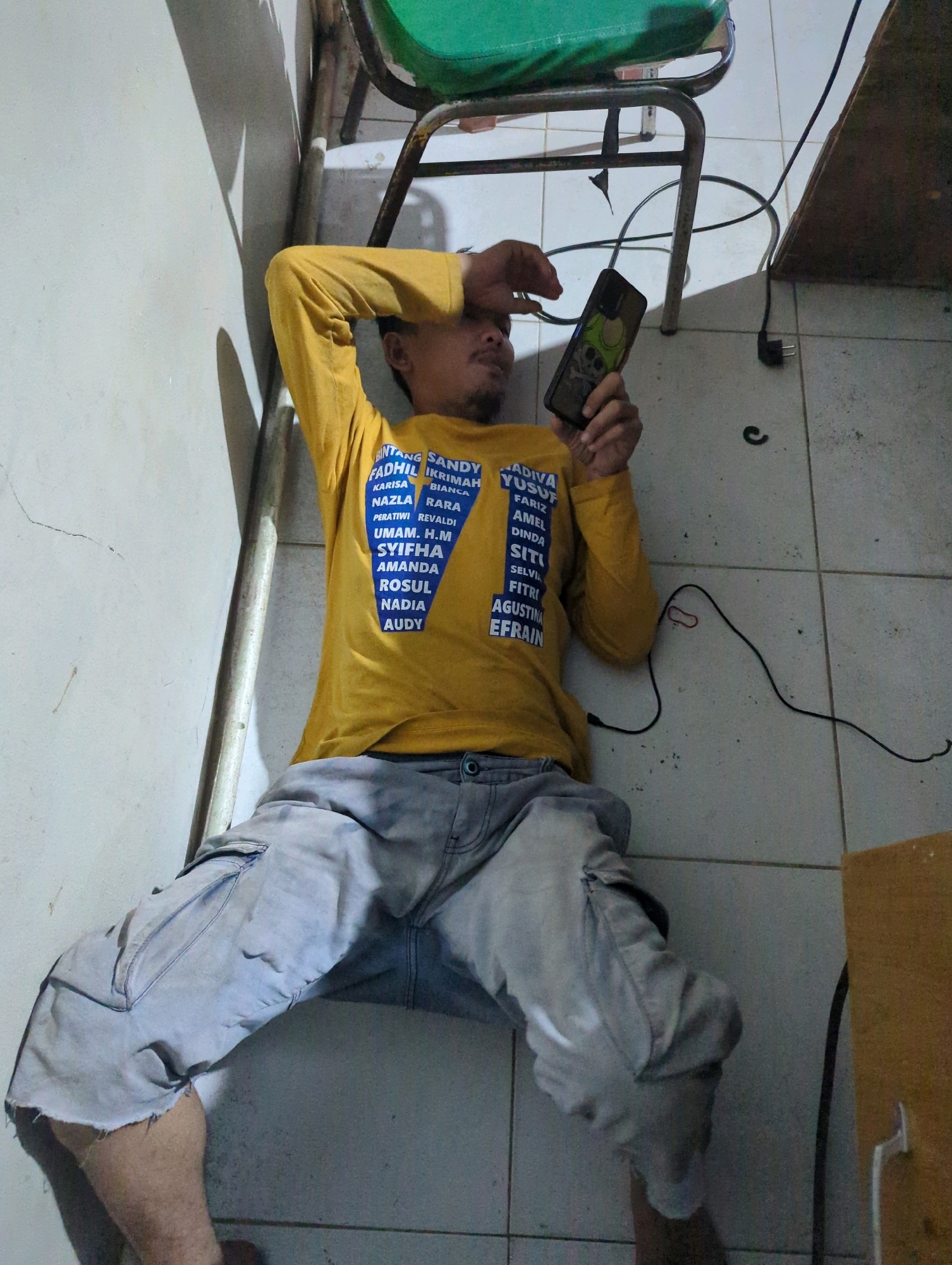 In Batu Belaman, Indonesia, a man is lying on his back on a tiled floor. He is wearing a yellow long-sleeved shirt with blue text and light grey cargo pants with torn hems. The man is holding a smartphone in his right hand, which is positioned over his forehead. A metal chair with a green seat is visible behind him. Electrical cords and a power plug are on the floor near him. The environment appears to be indoors, possibly a room or hallway, with a textured wall to the left and a wooden object to the right.