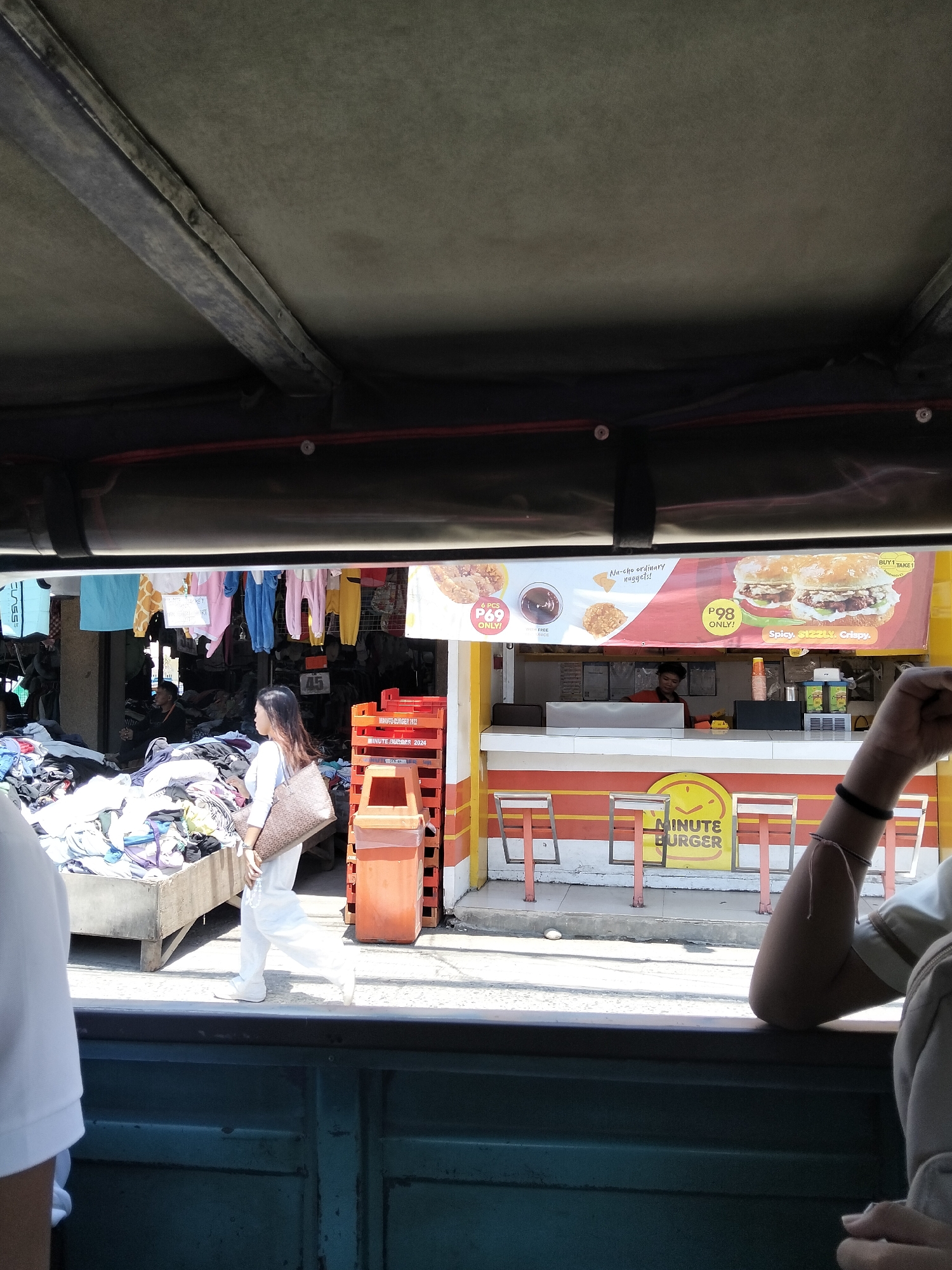 The image captures a street scene in Davao City, Philippines, viewed from inside a vehicle. The foreground shows the interior of the vehicle with a dark green painted metal surface. In the middle ground, a woman in a white outfit and a patterned bag walks past a stall. Behind her, a market stall displays a large pile of clothes. To her right, a fast-food stall with a bright yellow and red facade advertises "MINUTE BURGER." A banner above the stall features images of burgers, nuggets, and pricing, with text reading "BUY 1 TAKE 1," "P69 ONLY!," and "P98 ONLY!." The scene appears to be set during the daytime, with bright sunlight casting shadows. The atmosphere seems to be that of a bustling market or commercial area. Visible text includes brand names, prices, and promotional messages related to food items.