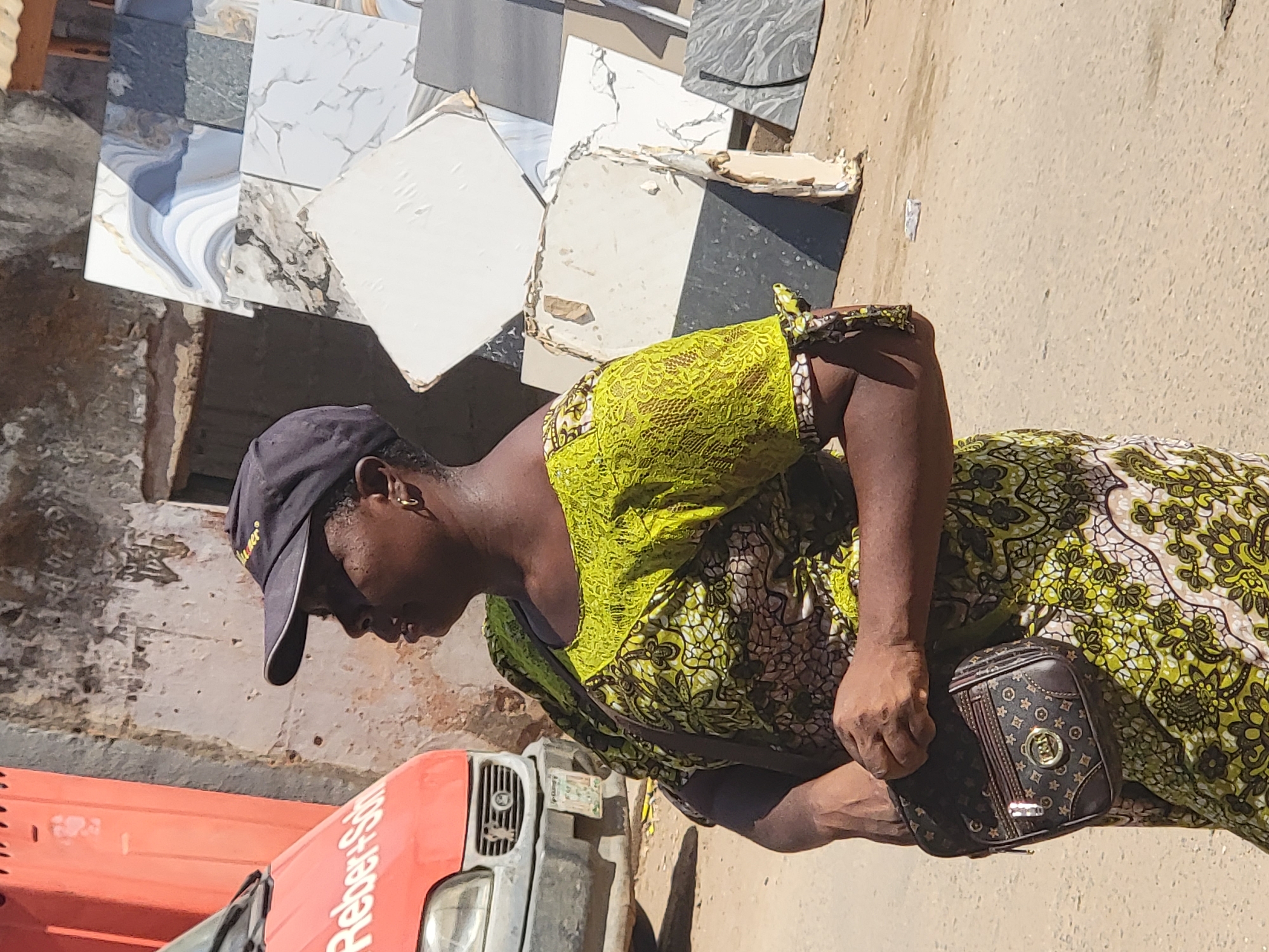 A woman in Maiburiji, Nigeria, wearing a patterned green and yellow dress and a dark baseball cap is walking outdoors on a sunny day. She carries a small brown patterned purse. Adjacent to her is a red vehicle. Behind her are stacks of tile.