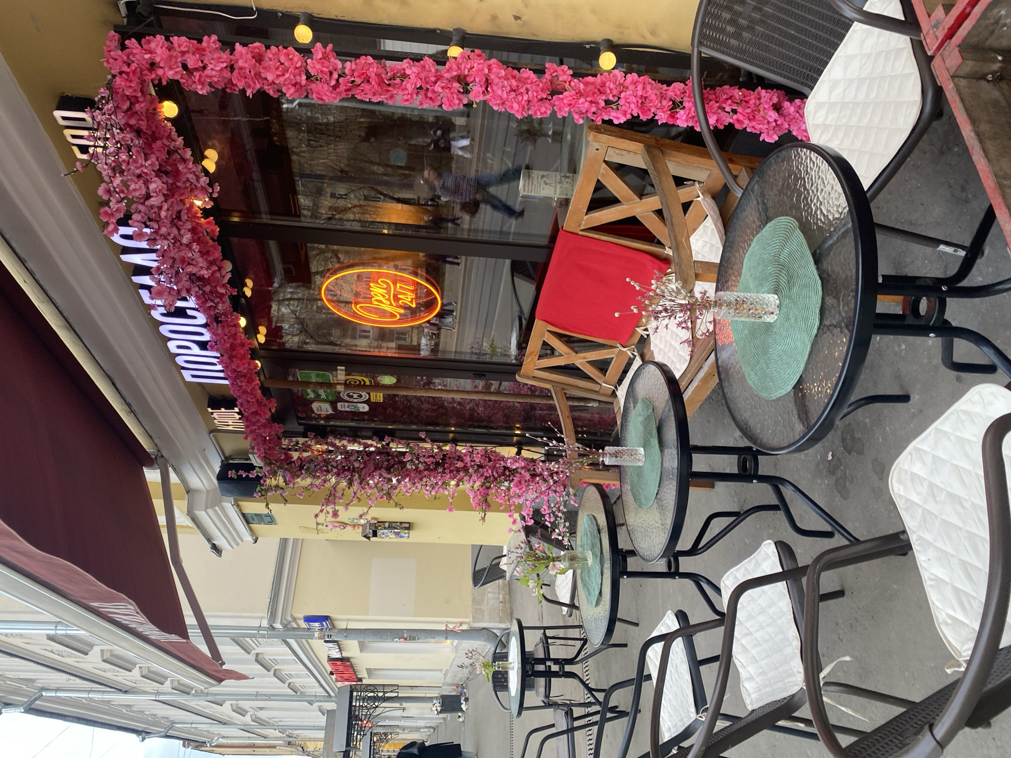 The image shows an outdoor cafe scene in Moscow, Russia. The cafe has tables and chairs set up outside, decorated with pink floral garlands. A neon sign reading "Open 24/7" is visible in the cafe window. The cafe's awning is a dark burgundy color. People are reflected in the cafe window.