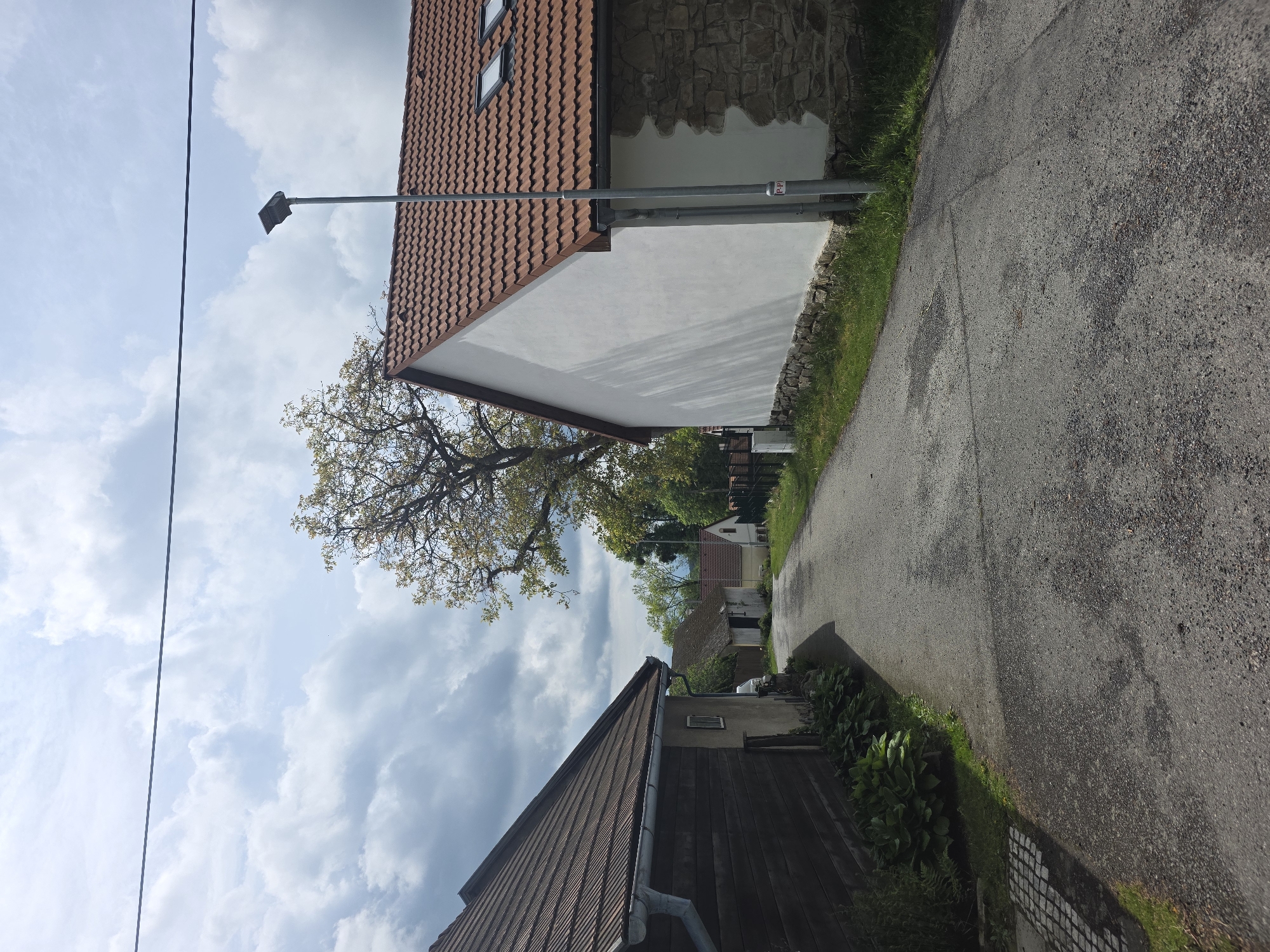 The image depicts a narrow asphalt road in an unidentified location within the city of Unknown, Unknown. The road is flanked by buildings with tiled roofs and a building with a combination of stone and white-painted walls. Vegetation lines the edges of the road, and a tree with sparse foliage is visible. A street lamp is positioned near the building with the tiled roof. Power lines are strung overhead against a cloudy sky.