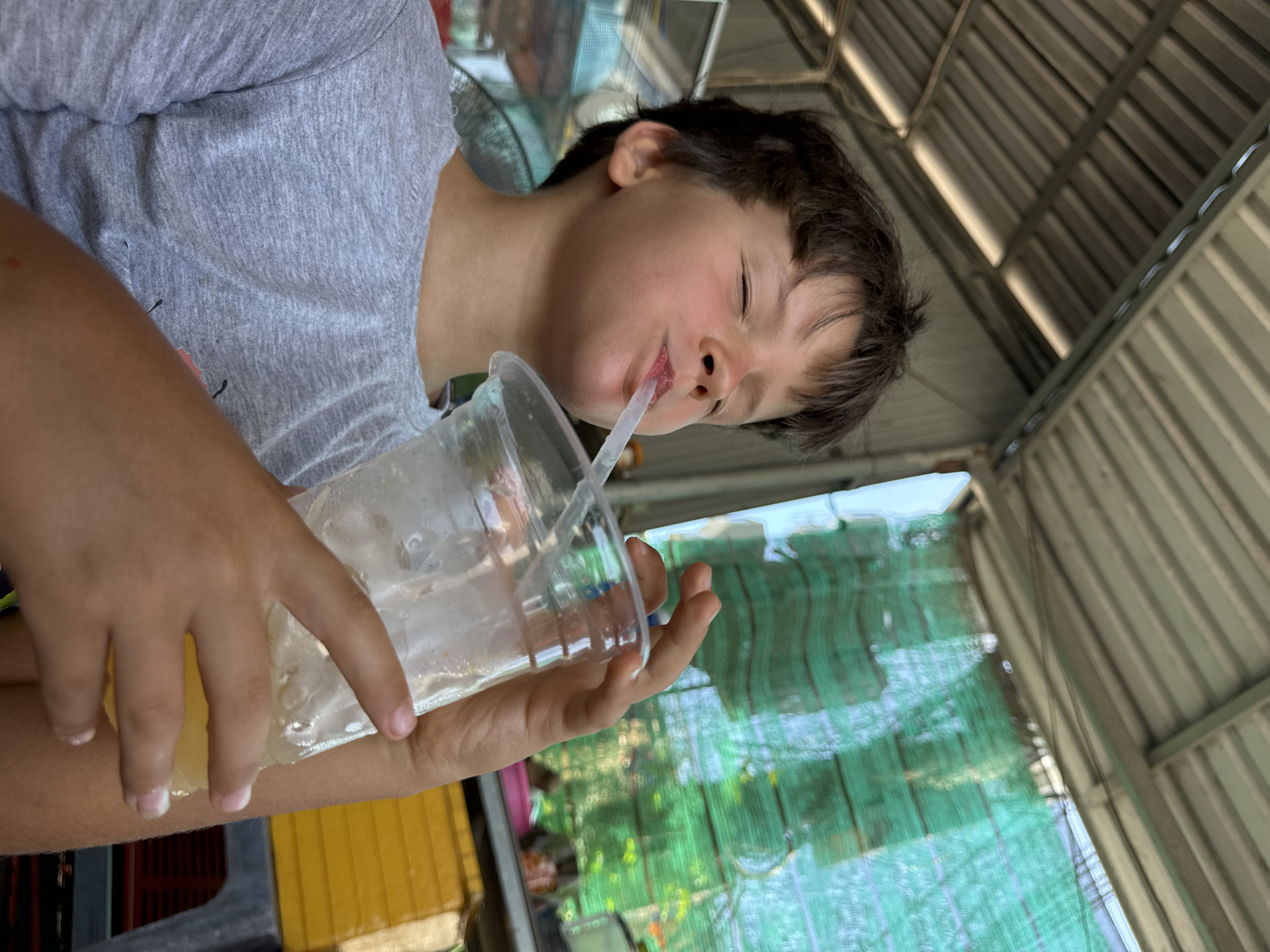 A young person is drinking from a clear plastic cup with a straw. The person is wearing a grey t-shirt. They are in an outdoor setting, possibly a market or roadside stall, with a corrugated metal roof overhead and green netting in the background. The lighting suggests it is daytime. The person's eyes are closed, and their face is tilted back as they drink, suggesting a moment of enjoyment or relaxation. Their hand is visible holding the cup. The overall scene is casual and appears to be a candid shot. There are some yellow crates visible to the left of the frame.