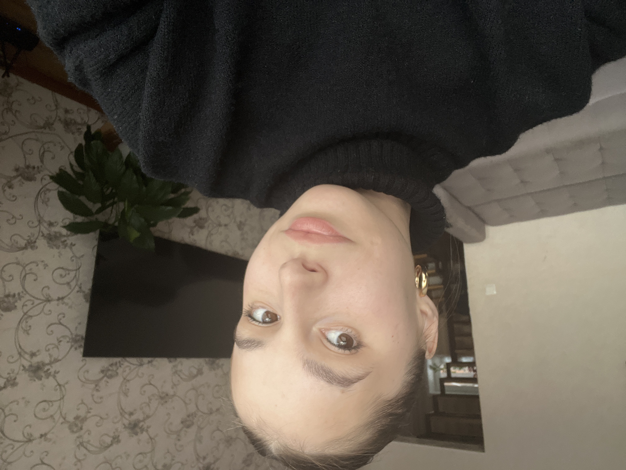 The image, when correctly oriented, features a young woman looking directly at the camera. She has brown eyes, a neutral expression, and light skin, with her dark hair pulled back. She is wearing a black turtleneck sweater and a gold hoop earring on her visible left ear.
The setting appears to be an indoor domestic space, likely a living room, located in Chaikovsky, Russia. The background on the left displays patterned wallpaper with a grey and white floral or damask design. Behind her, to the left, a flat-screen television and a green potted plant are visible. To the right of the woman, a light grey upholstered sofa or armchair is partially seen, alongside a plain, light-colored wall.
The lighting suggests it is daytime, providing soft, natural illumination. The primary activity is the woman posing for the photograph. A subtle reflection in a surface near her right ear shows what might be stairs or shelves in another part of the room. No visible text is present in the image.
