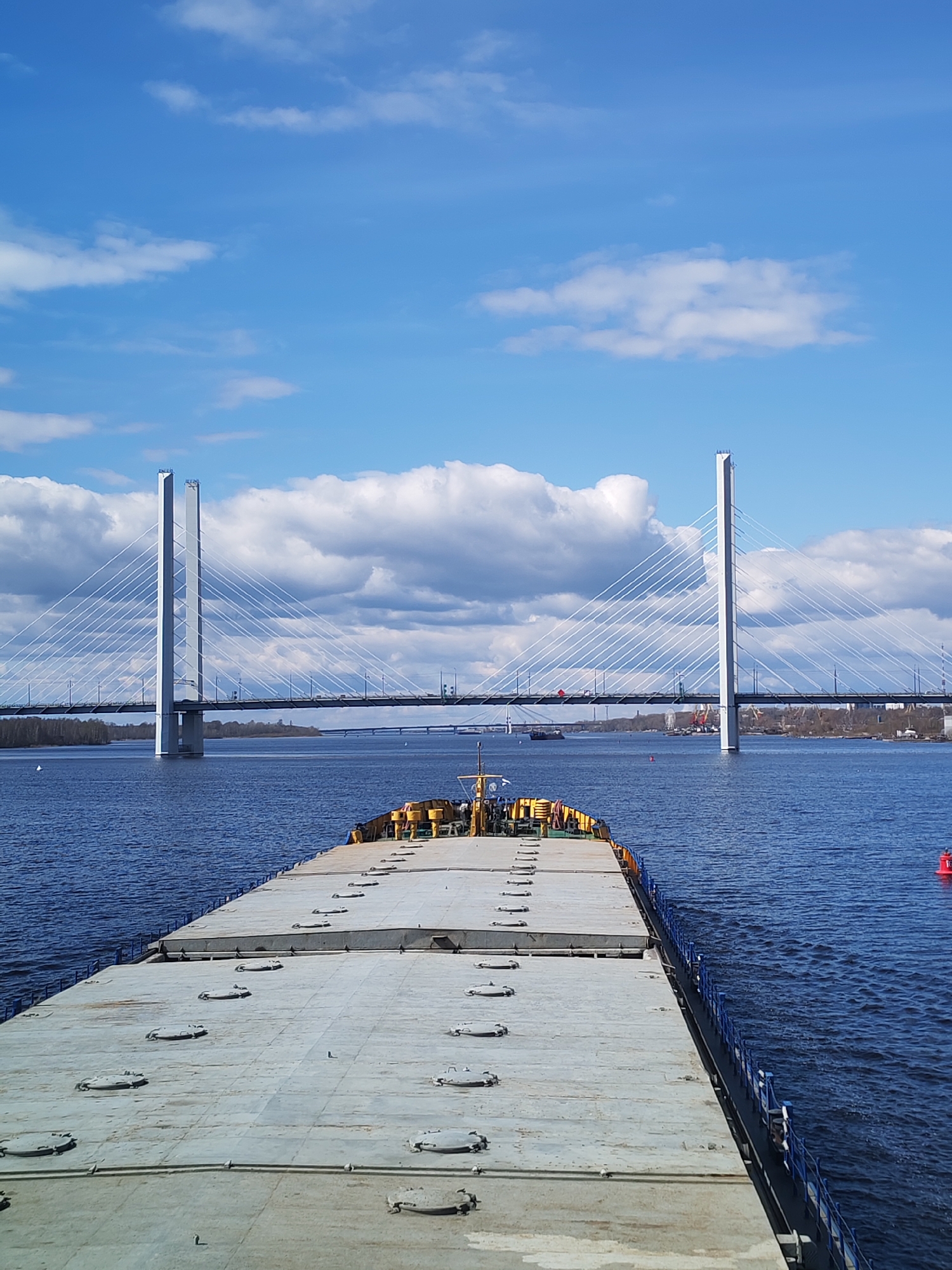 The image presents a daytime scene from the deck of a large vessel, likely a barge or cargo ship, navigating a wide body of water towards a prominent cable-stayed bridge.
In the foreground, the flat, light-grey deck of the vessel dominates the lower portion of the frame. It features multiple circular metal hatches, evenly spaced across its surface. Towards the bow, some yellow equipment or structures are visible. A blue railing runs along the starboard side of the vessel. To the right of the vessel, a red cylindrical buoy floats in the blue water, which shows gentle ripples.
The midground is dominated by a modern cable-stayed bridge. It has two tall, slender, light-colored pylons from which numerous white cables extend downwards, supporting the bridge's roadway. Vehicles are visible on the bridge deck, indicating it is a road bridge. Further in the background and to the right of the main bridge, a lower, older-looking bridge can be seen spanning the water. The riverbanks feature green foliage from trees and faint outlines of distant structures, suggesting either urban or industrial development. A smaller, dark vessel is visible on the far-left side of the river, underneath the main bridge.
The sky above is bright blue with scattered white cumulus clouds, indicating fair and sunny weather. The light suggests it is either morning or early afternoon. The overall scene conveys a sense of calm and smooth passage along the waterway. No discernible text is visible anywhere in the image.