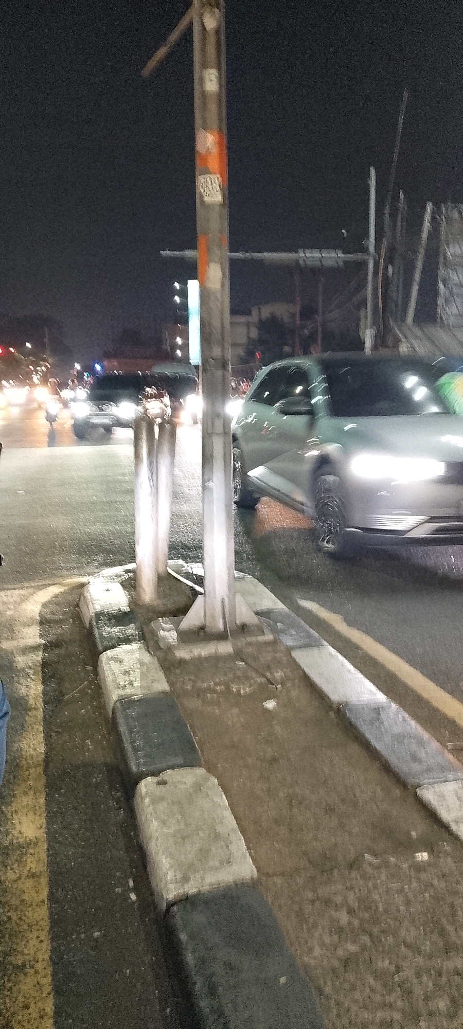 The image depicts a street scene at night in Depok, Indonesia. A silver car with its headlights on is visible in the foreground, positioned on the road. The car appears to be moving or has recently stopped, with motion blur evident. Adjacent to the road, there is a sidewalk area with a concrete curb made of alternating black and gray rectangular blocks. A metal pole, possibly a street lamp or traffic signal support, stands on the sidewalk near the curb. The pole has several stickers on it, including some with orange accents and text. In the background, other vehicles with illuminated headlights and taillights are present on the road, indicating traffic. The overall environment suggests a city street at night, with artificial lighting from vehicles and possibly streetlights. The ground appears wet, indicating recent rain or a damp surface. There are no clearly identifiable people in sharp focus, though blurred figures might be present in the background.