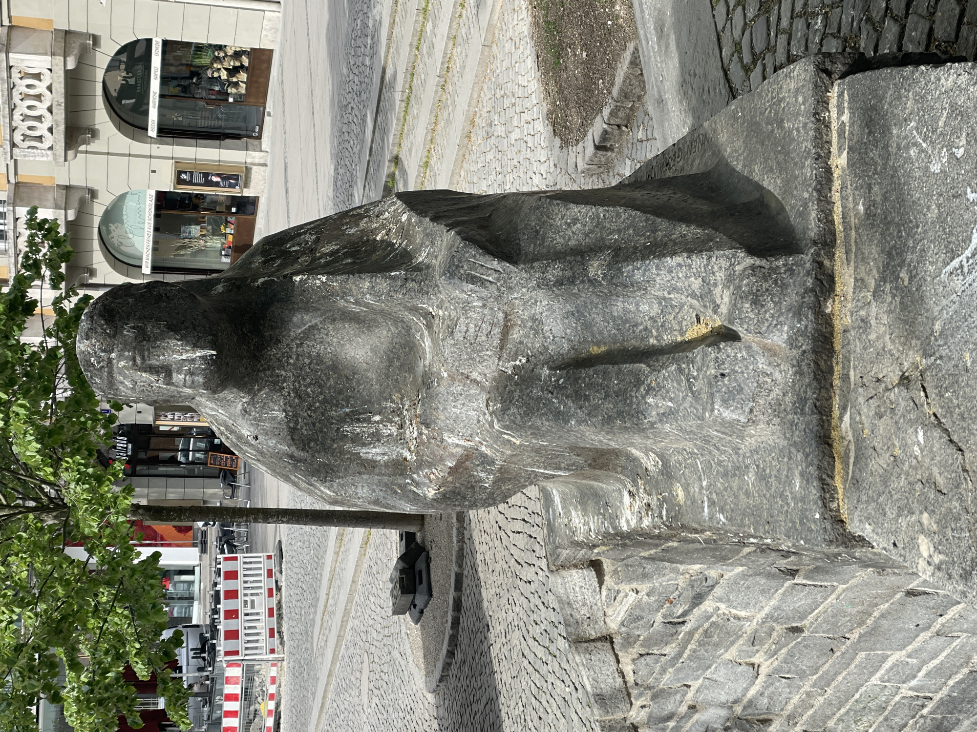 A wide-angle, vertically oriented photograph (likely rotated from a landscape capture) depicts a large, abstract stone sculpture set in an urban environment under bright daylight. The sculpture, crafted from dark grey, rough-hewn stone, suggests a stylized human form, possibly seated or reclining, and dominates the right side of the frame.
The ground surrounding the sculpture is paved with grey cobblestones. In the background to the left, architectural elements of multi-story buildings are visible, including arched windows and doorways. Prominent text on a shop window reads "HAARSTUDIO" at the top, with "HAARSTYLING," "EXTENSIONS," and "FRISEUR" also discernible. Below this, another shop displays "WIR MACHEN FEINES AUS SCHOKOLADE," indicating a chocolate shop.
Further in the background and to the right, a tree with lush green leaves stands alongside red and white striped barriers, commonly used for construction or cordoning off areas. More paved surfaces and other distant buildings can be seen. The scene is illuminated by strong sunlight, casting distinct shadows, indicating a clear, sunny day. No people are visible, and the scene is static, focusing on the sculpture and its urban context. The sculpture shows varying textures and some lighter discolorations, possibly from weathering or deposits.