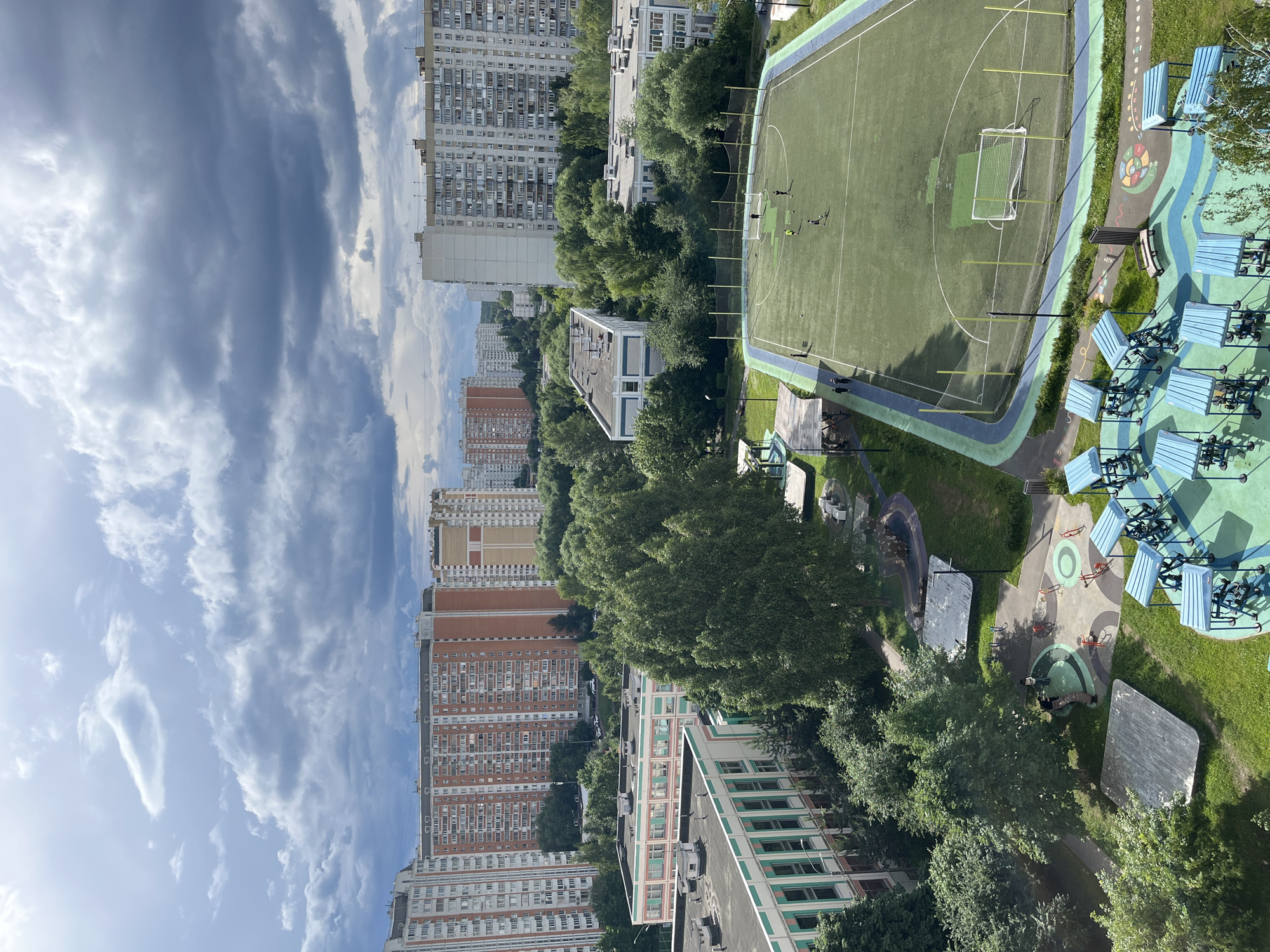 This is an aerial view of an urban residential area in Moscow, Russia, on a cloudy day, likely in the late morning or early afternoon given the brightness of the sun even through the clouds.
The scene features a sports field with green turf, marked with white lines and a soccer goal. Several individuals are scattered across the field, appearing to be playing or practicing. Adjacent to the field is a children's play area with colorful, patterned ground surfaces, benches, and some playground equipment. A paved pathway winds through the area.
Surrounding the sports and play facilities are several multi-story apartment buildings of varying architectural styles and colors. Some are light-colored with numerous windows, while others are brick-red with balconies. Dense green trees are interspersed throughout the residential blocks and around the recreational areas, indicating a well-established neighborhood. The sky is filled with dramatic, textured clouds, suggesting changeable weather. The overall impression is of a developed urban environment with ample green spaces and recreational facilities for residents. No visible text is present in the image.