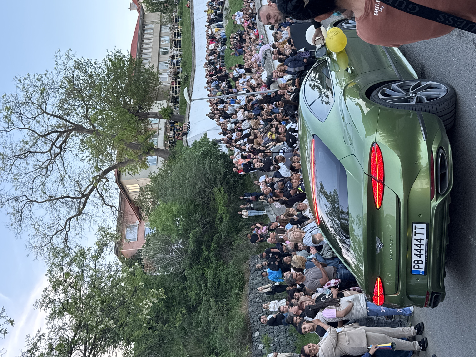 Captured from a high vantage point, the image presents a scene likely unfolding on an incline in Tsarevo, Bulgaria. A large crowd has gathered, filling both an elevated terrace and a lower area fringed by greenery. The architecture indicates an urban setting with buildings visible on the upper level. The crowd's attention is focused away from the camera, possibly towards a stage or focal point not directly visible. In the immediate foreground stands an olive-green Bentley, positioned as a prominent element within the scene, with a yellow balloon tethered to the side mirror. The vehicle's license plate reads "PB 4444 TT" and bears the Bulgarian flag. A person partially obscures the upper-right corner, wearing a "SMILEY" labeled shirt and a crossbody bag marked "GUESS". The vegetation includes flowering trees and bushes, suggesting a temperate climate and contributing to the overall outdoor atmosphere.