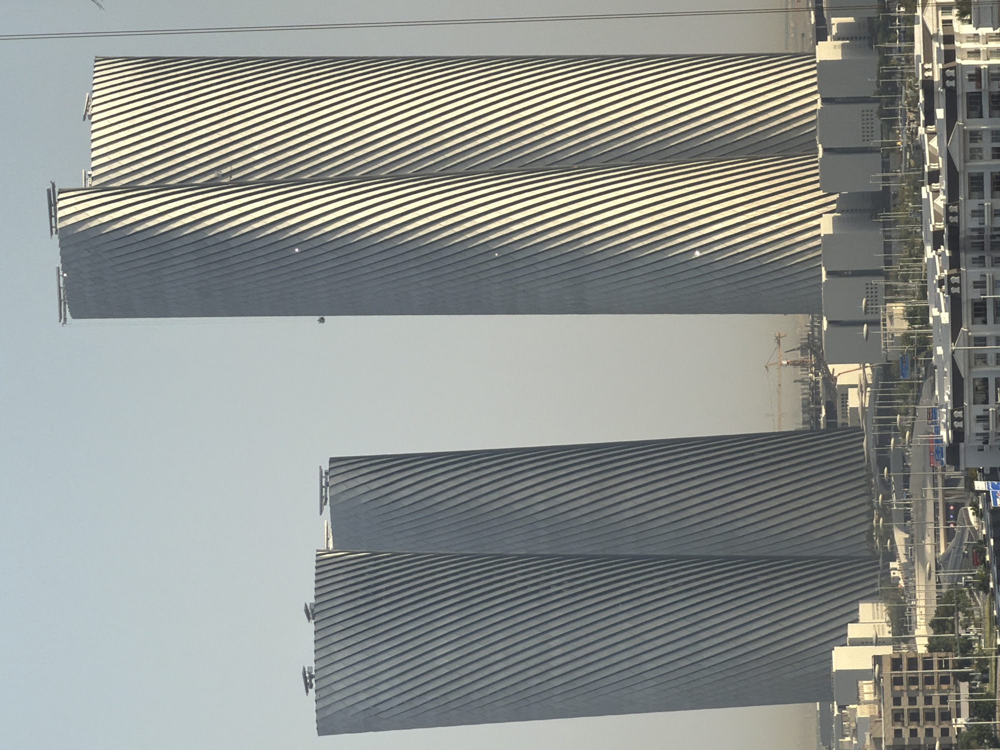 The image shows two strikingly tall, modern buildings with a unique twisted design. Their exteriors are clad in a material that reflects light, creating a ribbed effect with alternating dark and light stripes that spiral upwards. The building on the right is taller and appears to be composed of two distinct yet connected towers, while the one on the left is slightly shorter and also seems to be a paired structure. Both buildings have flat tops, with what appear to be some rooftop structures or equipment visible.
In the foreground and mid-ground, there is a cityscape with various other buildings of different architectural styles and heights. Some are low-rise structures with balconies and visible windows, while others are more modern, boxy buildings with flat roofs. There are also numerous streetlights and what appear to be traffic lights or signs visible on poles, suggesting a developed urban environment with infrastructure for transportation. A construction crane is visible in the distance to the left of the taller buildings, indicating ongoing development in the area. The overall atmosphere is bright, with a clear, pale sky, suggesting it is daytime. The lighting suggests the sun is positioned to create strong shadows on the twisted facades of the two main towers, highlighting their distinctive design.