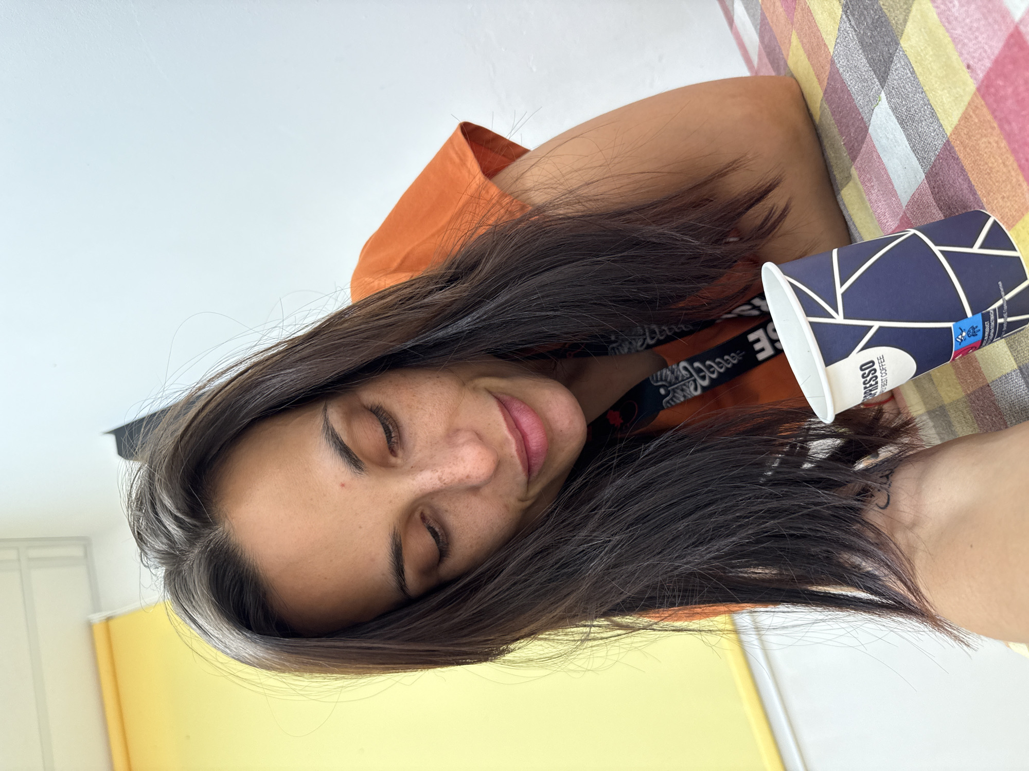 A woman with dark hair and freckles looks down at a coffee cup. She is wearing an orange t-shirt and appears to be indoors, possibly in a cafe or restaurant in Pleven, Bulgaria. The background is out of focus, but shows a yellow wall and a white wall. A checkered tablecloth is visible on the table in front of her, and the coffee cup has a blue and white geometric pattern. The scene is captured from a close-up, low-angle perspective.