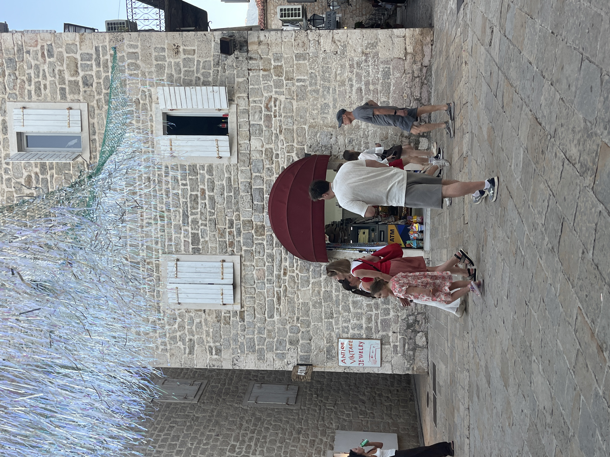 A street scene in Budva, Montenegro, features a stone building with an arched awning over a shop entrance. Several people are walking on the cobblestone street, including a woman in a red top and skirt, a young girl in a floral dress, and two men in casual attire. Overhead, a decoration of shimmering, iridescent strips hangs, adding a festive atmosphere. A sign on the building reads "ANTIQUE VINTAGE JEWELRY" and an ATM is visible. The architecture is typical of the old town, with stone walls and wooden shutters on the windows.
