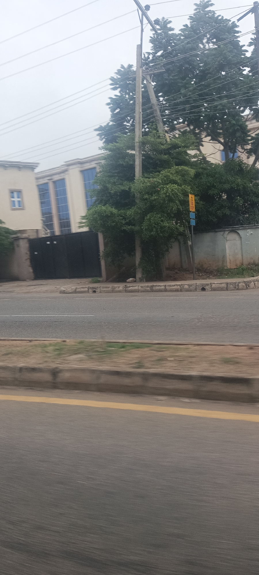 This is a daytime photograph capturing a street scene, likely taken from a moving vehicle, indicated by the motion blur on the lower half of the image. The setting appears to be a suburban or urban area in Jos, Nigeria.
In the foreground, a paved road with a distinct yellow line on the left and a fainter white line further into the lane is visible. A concrete curb separates the road from a narrow strip of dirt and sparse grass, which then leads to a sidewalk-like area with concrete blocks.
The mid-ground features dense green foliage from trees and bushes, partially obscuring a building. A tall utility pole with multiple overhead wires stands prominently on the right side of the frame, emerging from behind the greenery. To the right of the utility pole, a yellow and blue sign is visible, marking a "BUS STOP" area. Behind the bus stop sign, there's a light-colored wall with an arched decorative element.
In the background, a light-colored building with multiple tall, blue-tinted windows is visible. A dark, solid fence or gate is in front of this building. The sky is overcast or hazy, providing soft, diffused lighting. No people or animals are visible in the frame, and no specific activities are taking place other than the presumed movement of the camera's platform. The architecture and street elements are consistent with an urban environment in Nigeria.
