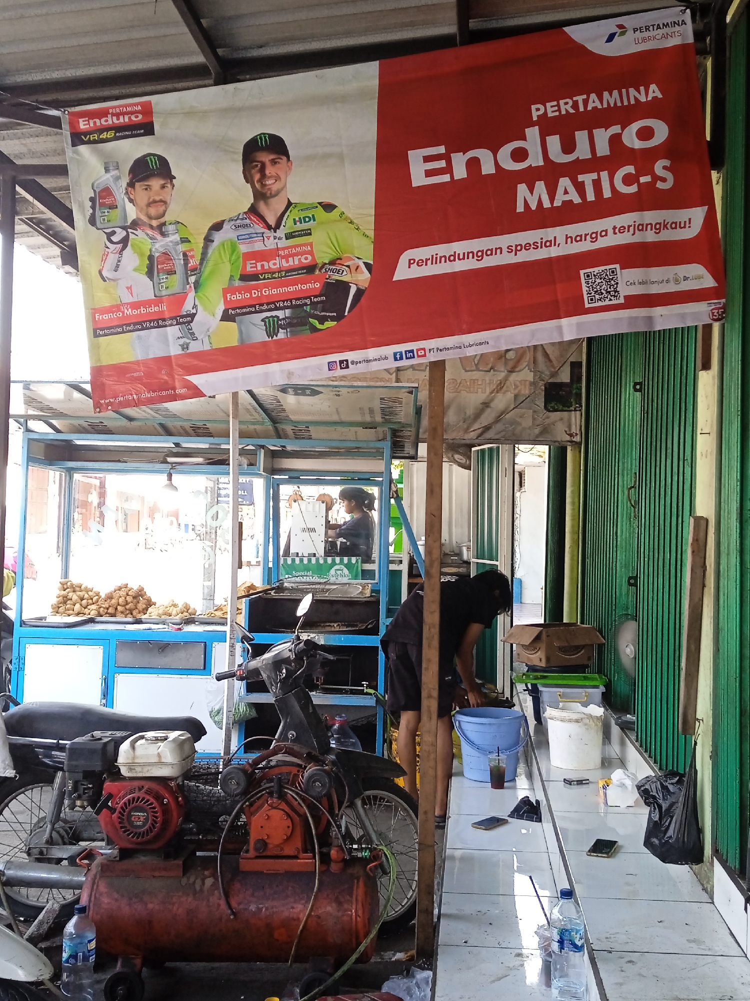 The image shows a street food vendor in what appears to be Pandean, Indonesia, based on the writing on the banners. A man is bent over, attending to something at the counter, with supplies like buckets and bottles of water around him. Behind him is a green corrugated metal wall. A blue food cart sits to the left, with a display of fried goods. Hanging above the cart is a banner advertising Pertamina Enduro VR46 Racing Team with Franco Morbidelli and Fabio Di Giannantonio. To the right is a banner for Pertamina Enduro MATIC-S oil, with Indonesian text and a QR code. A motorcycle and a red air compressor with a gasoline engine are parked near the food cart. Two smartphones lie on the tiled counter.