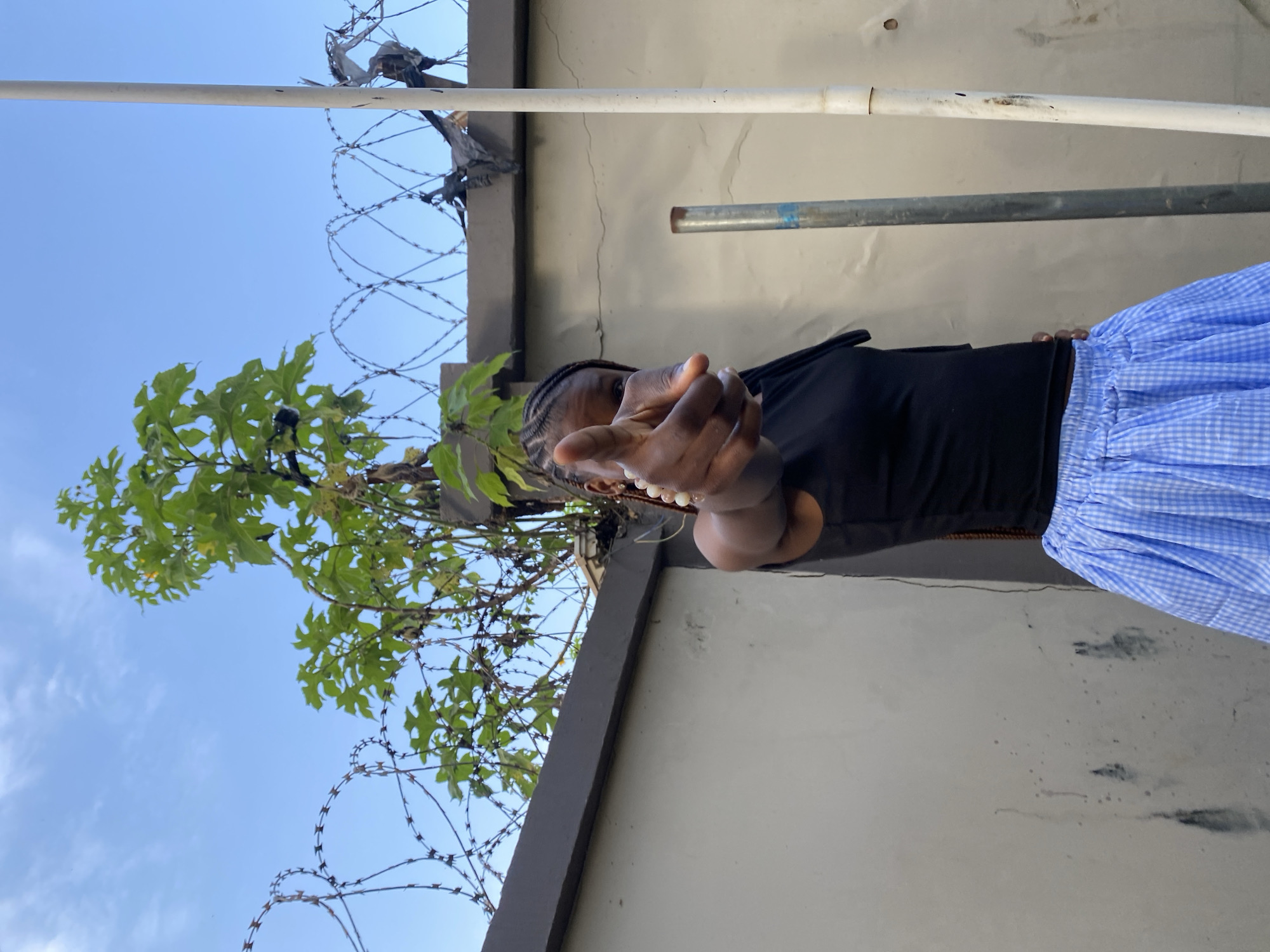 A young woman with braided hair, wearing a black tank top and a blue and white checkered skirt, points her index finger directly at the camera. She is standing in front of a light-colored wall, which is topped by a grey border fence. Barbed wire is strung along the top of the fence. A green plant with broad leaves grows from behind the fence, reaching towards the blue sky. Two poles, one metal and one white PVC, stand to the right of the woman. The scene appears to be an outdoor setting in Gura Top, Nigeria.
