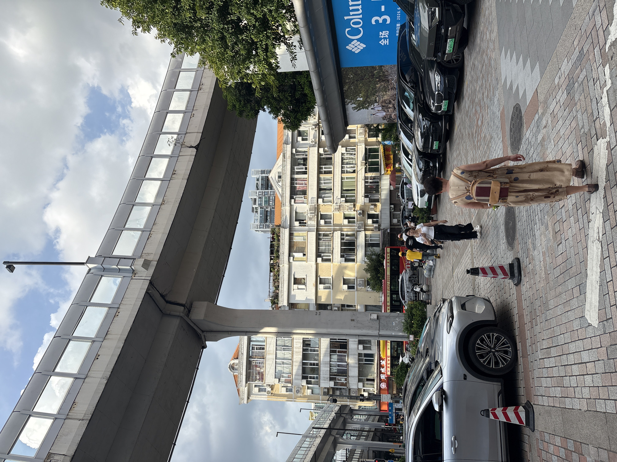The image depicts a street scene in Haizhu District, China, characterized by an elevated roadway structure dominating the upper portion of the frame. Below this structure, several buildings of varying architectural styles are visible, including multi-story residential buildings with numerous windows and balconies. The lower portion of the image shows a parking area with several cars, a sidewalk paved with interlocking bricks, and pedestrians walking.
In the foreground, a woman wearing a patterned dress and a backpack is walking away from the camera. To her left, a silver SUV is parked, with two striped traffic cones positioned in front of it. Further down the street, a red bus is seen, and other vehicles are parked. A large blue billboard with white text, possibly an advertisement, is attached to a building on the right side of the frame. The sky is partially cloudy, with patches of blue. The overall impression is of a bustling urban environment with a mix of modern infrastructure and older buildings.