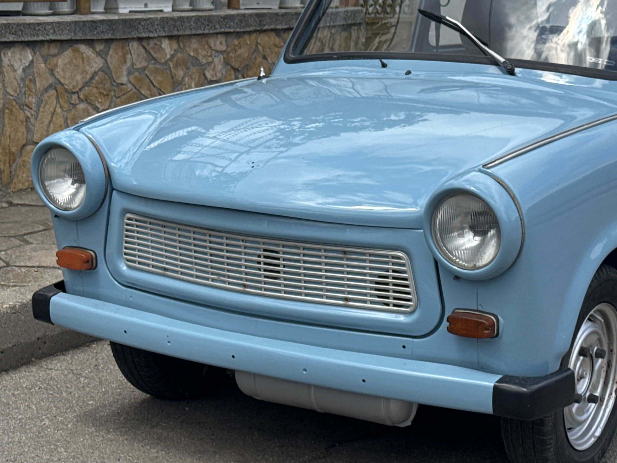 The image shows the front of a light blue Trabant car. It is parked on a street in an unknown location. The car is a two-door model, and the front features round headlights, a chrome grille, and a black bumper. The hood of the car reflects light from the environment, and the image is taken at eye level.