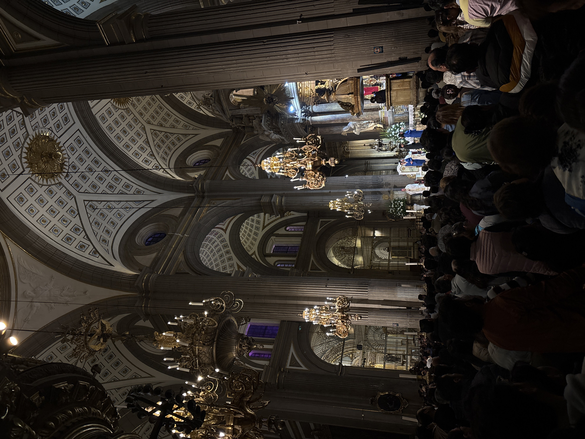 The image depicts a large crowd of people inside the Puebla Cathedral in Puebla, Mexico. The environment showcases the high ceilings with ornate designs, massive pillars, and golden chandeliers. People are standing and participating in a religious event near the altar. There are religious figures and floral arrangements around the altar area.