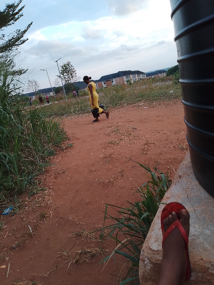 The image captures a scene in Ogboko, Nigeria. A woman wearing a yellow dress is walking on a red dirt path carrying a black bucket. In the background are buildings and street lights. In the foreground, the edge of a large water tank is visible, along with a foot in a red flip-flop. Grass and vegetation are also visible along the sides of the path.