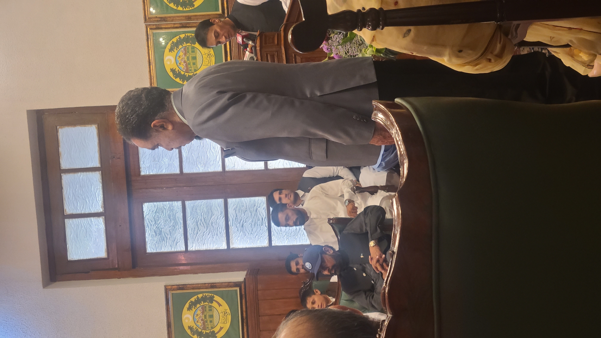A man in a gray suit stands near a window, with several other men seated behind him. The room is adorned with framed artwork. The location is in Quetta City Tehsil, Pakistan.
