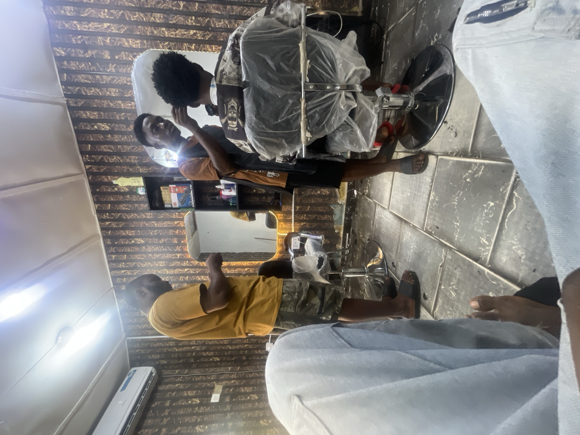 The scene appears to be inside a barbershop. There are two individuals visible. One, dressed in a brown shirt and camouflage shorts, is facing away from the camera and standing. The other, dressed in a dark shirt and shorts, is giving a haircut to an individual sitting in a barber's chair. The chair is partially covered in plastic. The barbershop has decorative wall panels and tiled floors. An air conditioning unit is visible. The location is Lokoja, Nigeria.