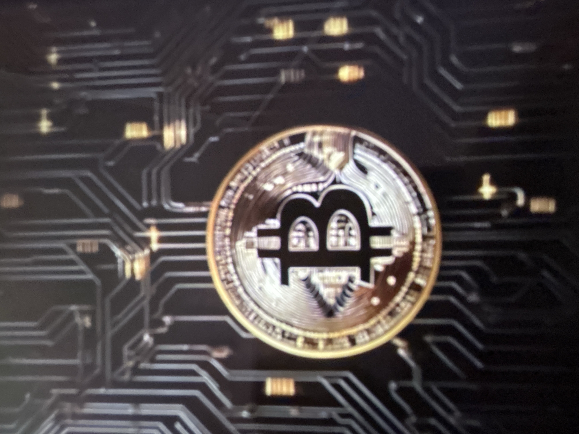 The image is a close-up shot of a Bitcoin cryptocurrency coin superimposed on a dark background that resembles a circuit board. The coin is golden and silver with the Bitcoin symbol prominently displayed. The circuit board has intricate lines and connections, with some areas appearing lit or reflective, suggesting a digital or technological theme. The lighting highlights the metallic sheen of the coin and the reflective surfaces on the circuit board. The image does not depict any specific location in Gelendzhik, Russia, or any people, actions, or interactions. It is a still, conceptual image.