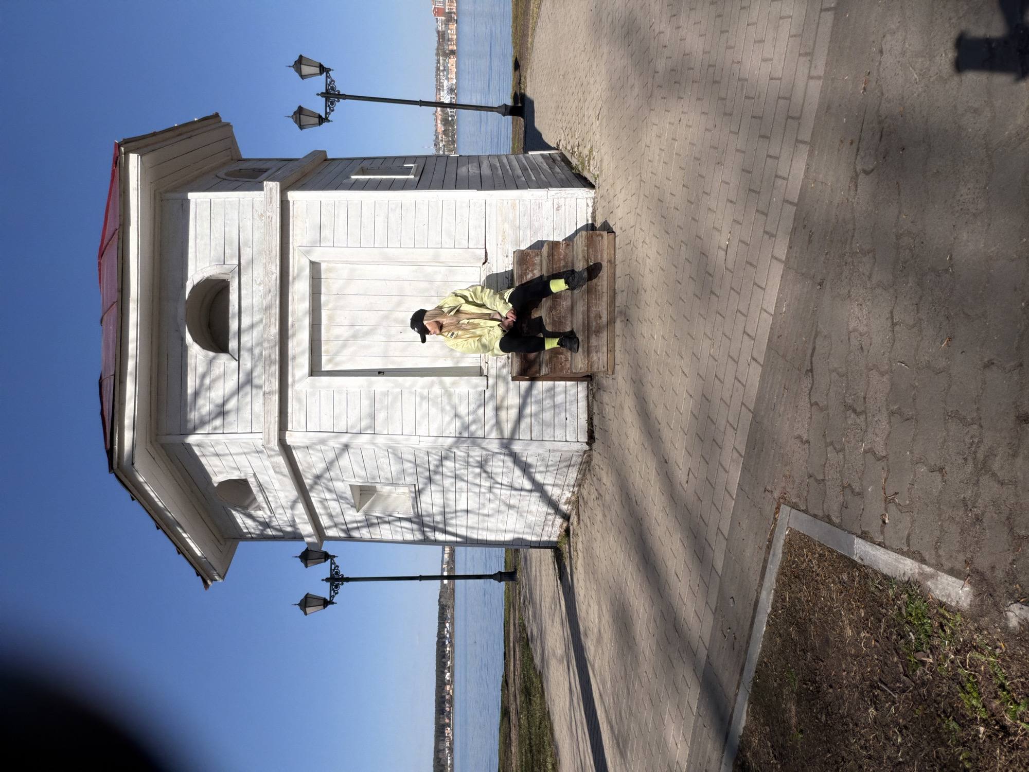 The image depicts a woman sitting on the steps of a small, white, wooden building with a red roof. The building is located on a paved pathway near a body of water, likely a river or lake. The pathway is lined with lamp posts. The water appears calm. Across the water, a shoreline with buildings can be seen. The woman is wearing a light green jacket, black pants, and a black cap. She is seated casually, with her legs extended. The location is identifiable as Votkinsk, Russia, due to the distinct architectural style of the building, which is a known landmark in the city. The day appears sunny, casting shadows from the building and trees.